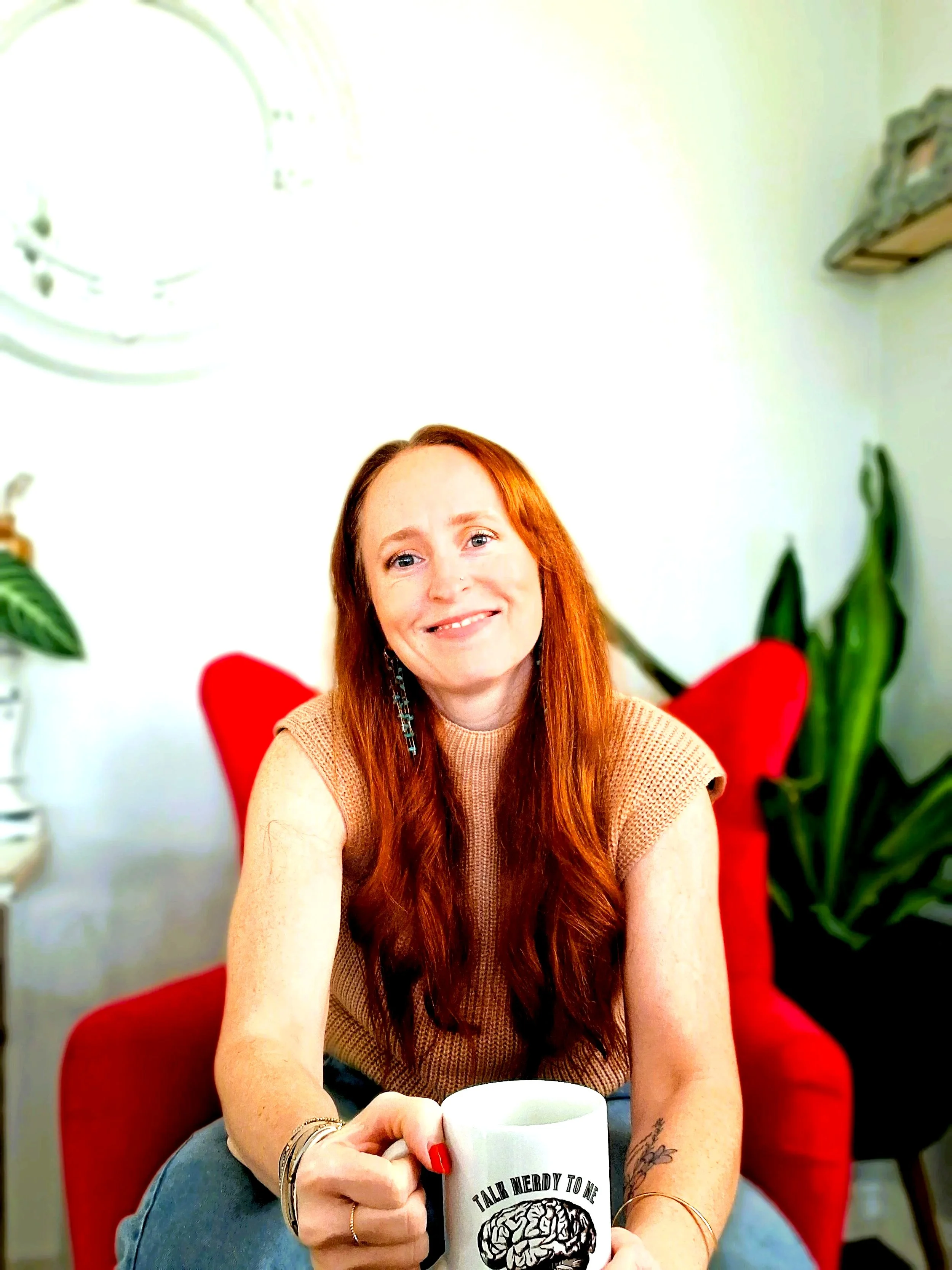 A woman with long red hair sitting in a chair, smiling, holding a white mug with a brain illustration and text, in a cozy room with plants and a white wall.