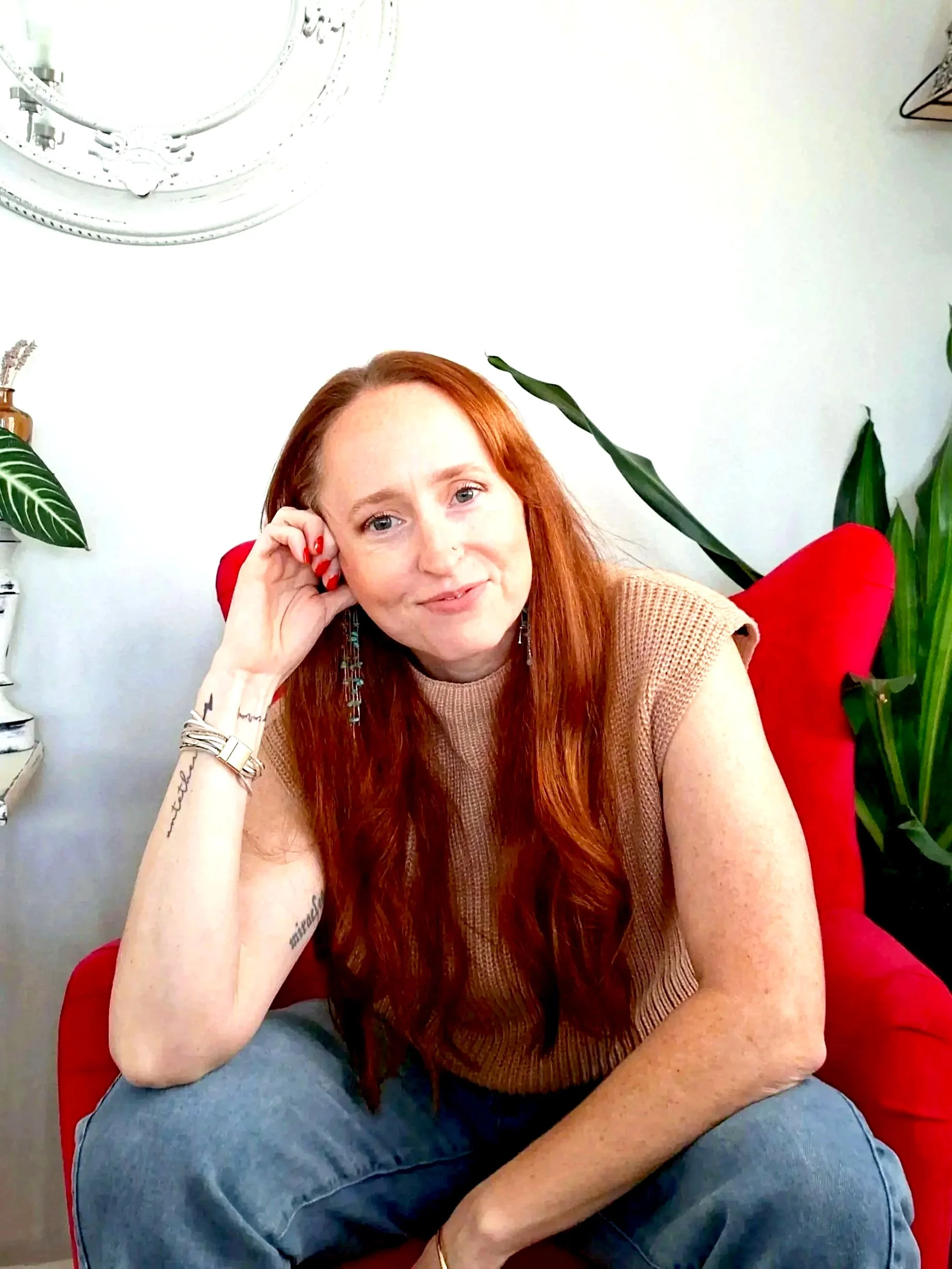 A woman with long red hair and light skin sitting on a red chair, leaning on her left hand, smiling slightly at the camera, with plants and a white wall in the background.