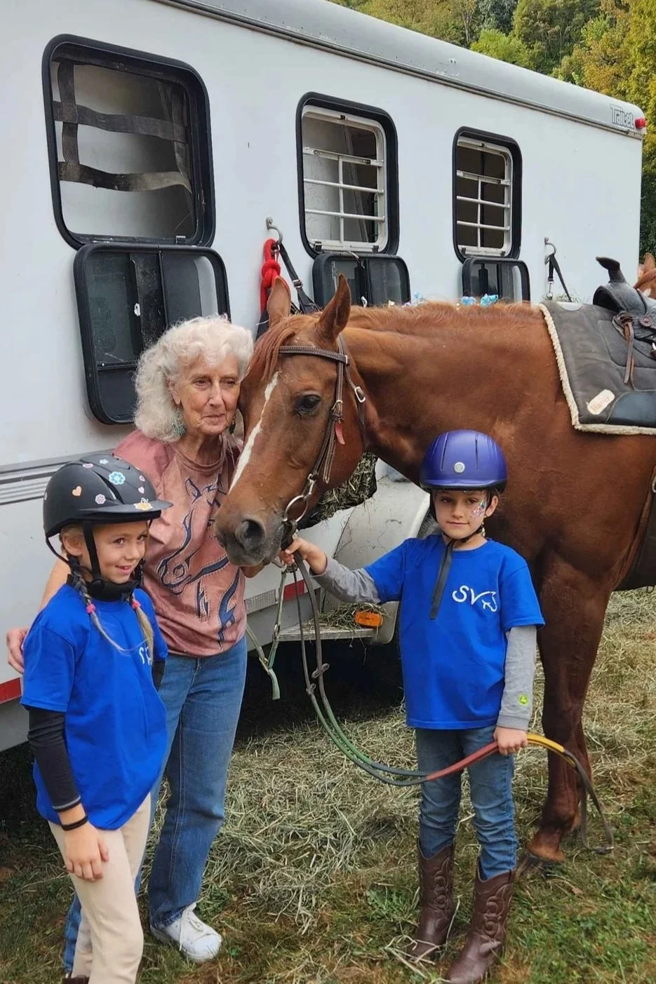 An elderly woman and two children standing next to a horse in front of a white horse trailer. The woman is petting the horse, and the children are wearing riding helmets and blue shirts. The surroundings suggest an outdoor setting.