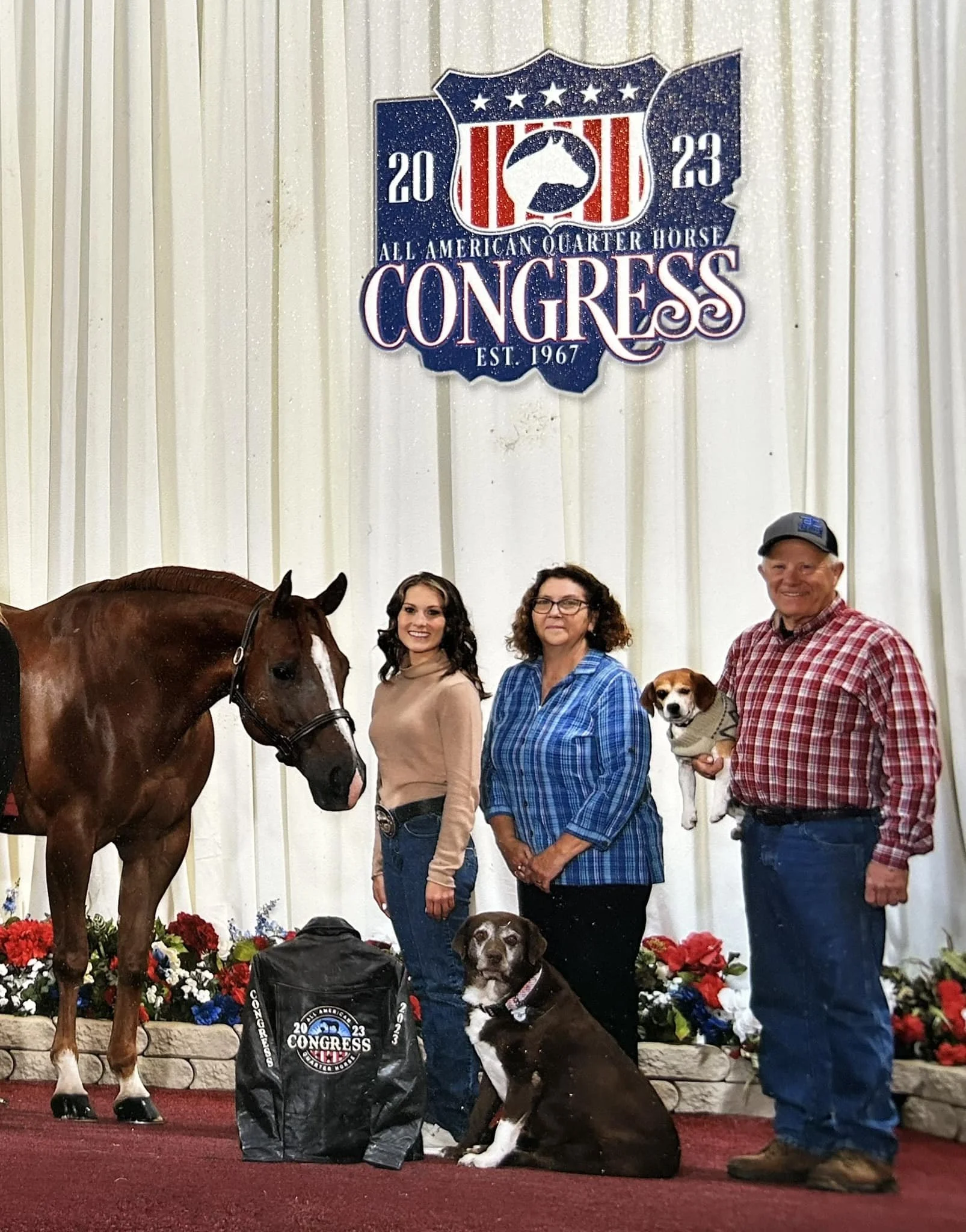 Group of four people, two women, one man, and one teenage girl, standing indoors in front of a white curtain backdrop with a sign that reads "All American Quarter Horse Congress, EST 1967" and features a horse head logo. They are accompanied by a large brown horse, a brown and white dog sitting on the floor, and a man holding a small dog. There is a black Congress-themed bag on the floor in front of the group with red, white, and blue flowers in the background.