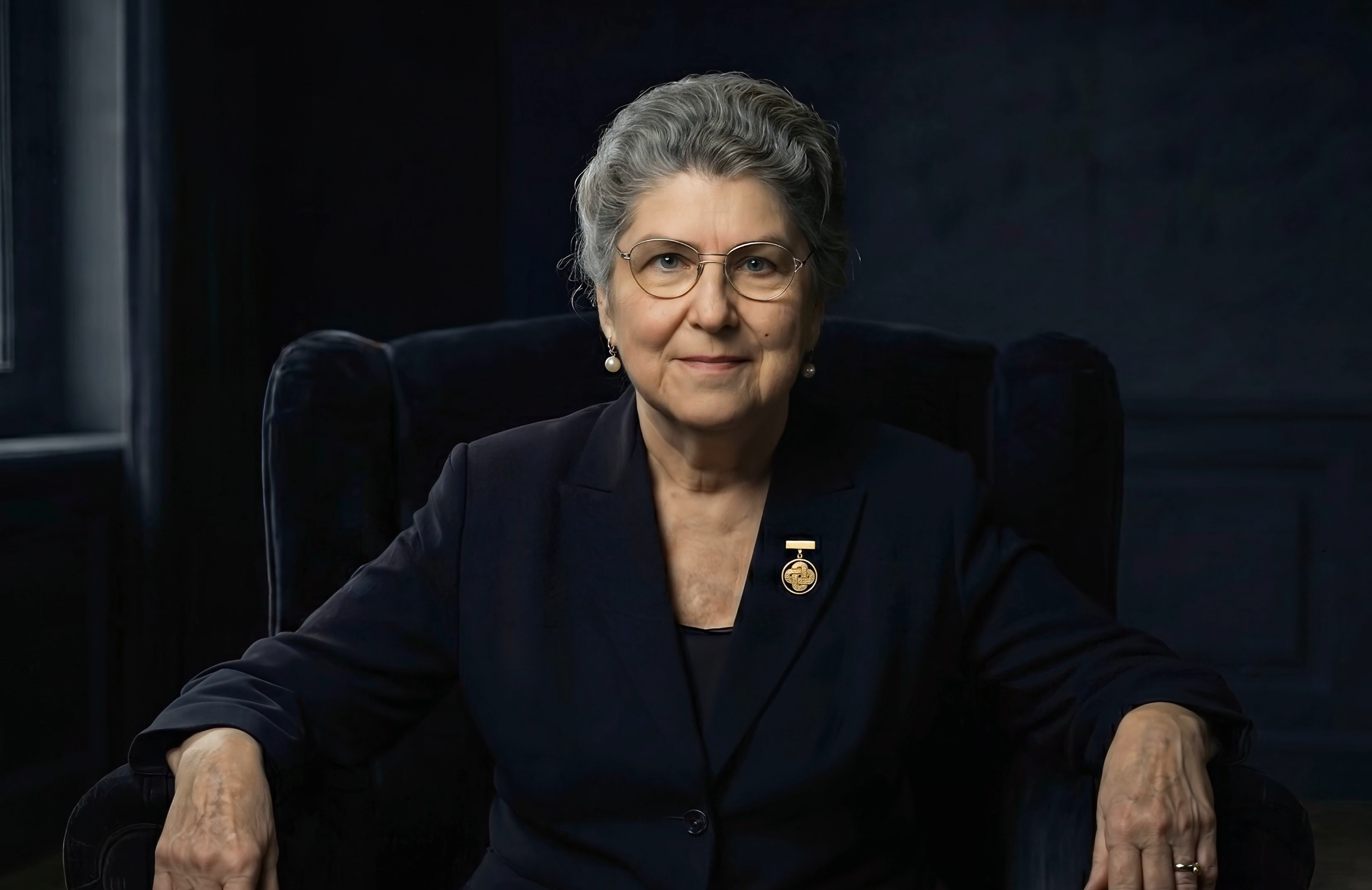 A portrait of an elderly woman with gray hair, glasses, and earrings, sitting in a dark room, wearing a dark blazer with a gold pin.