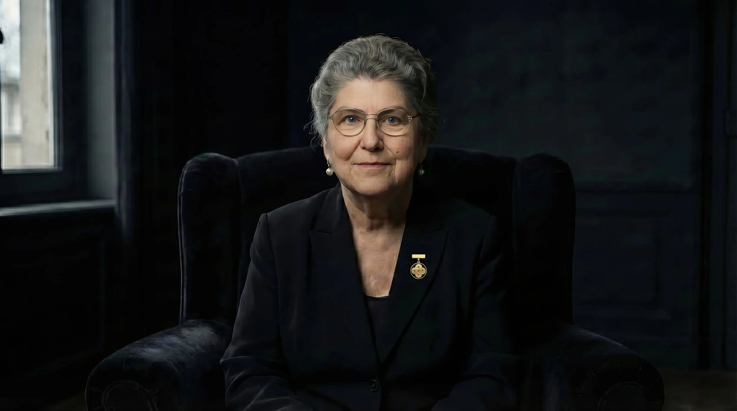 Marie de Caba, an elderly woman with gray hair, glasses, and pearl earrings sitting in a dark room with a black background. She is dressed in a black jacket with a gold brooch and pin, looking directly at the camera.