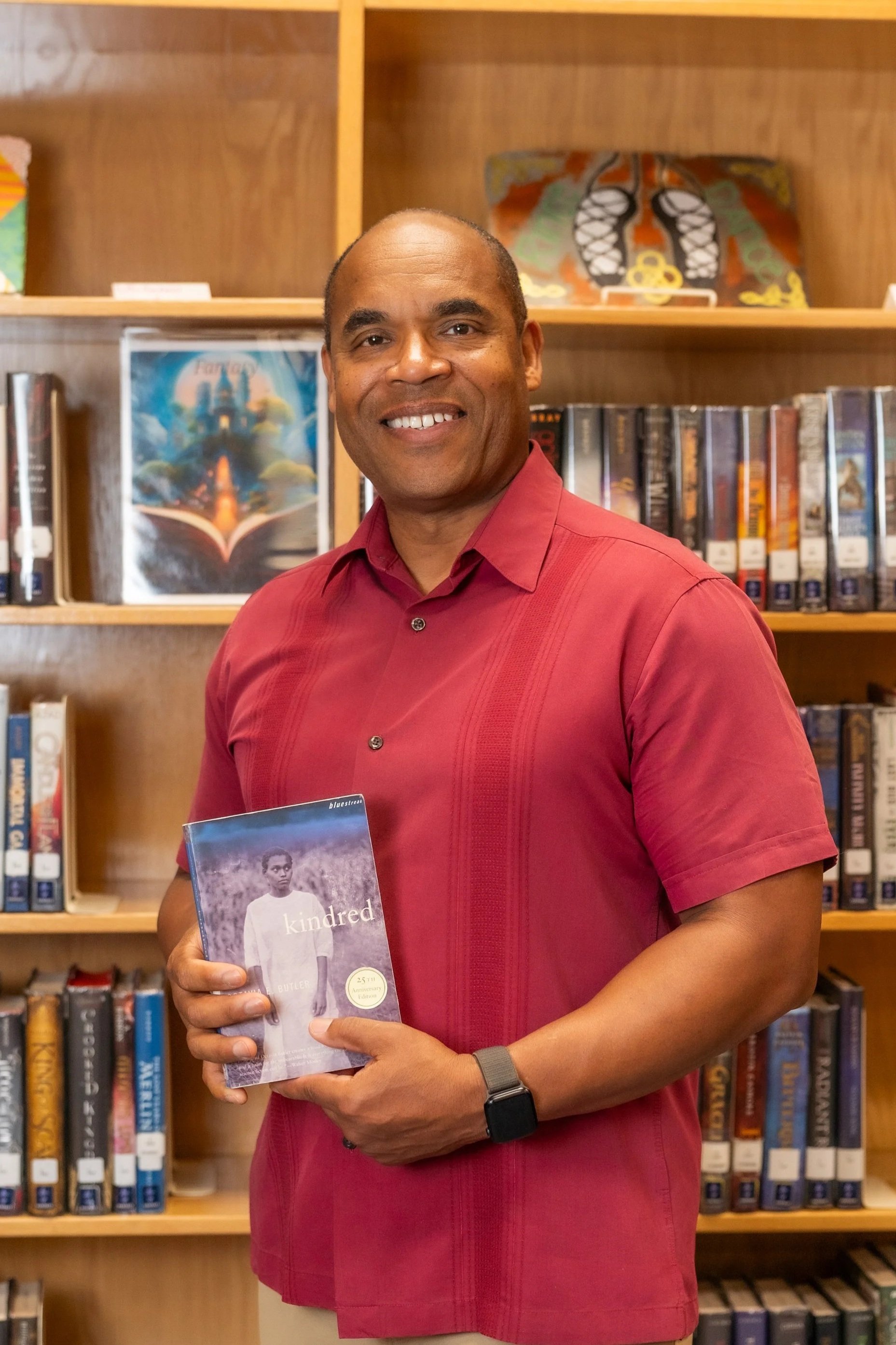 Mark Covington standing in front of a bookshelf holding a copy of "Kindred" by Octavia Bulter