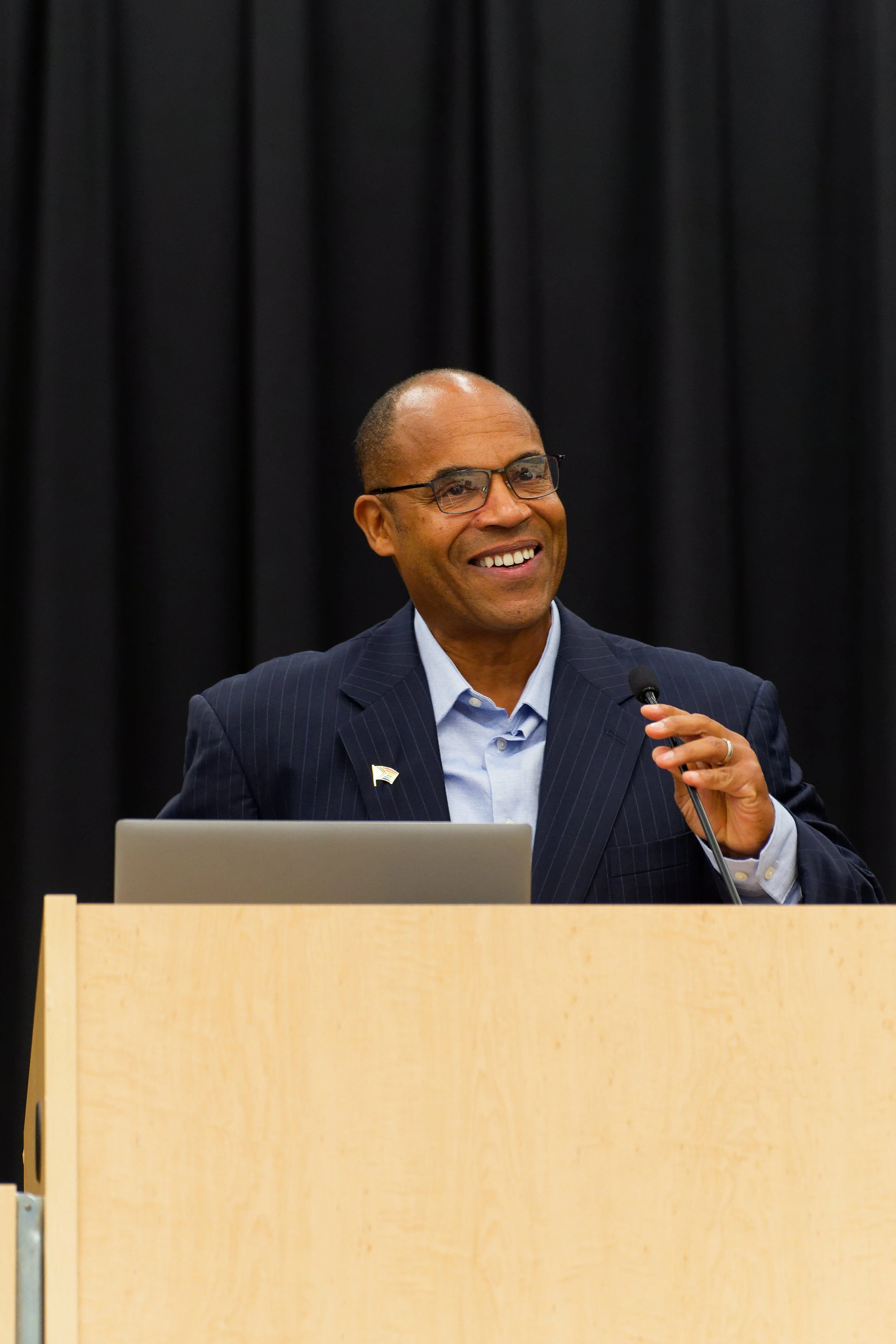 Mark Covington standing behind a wooden podium with a laptop and microphone in front of black curtains.