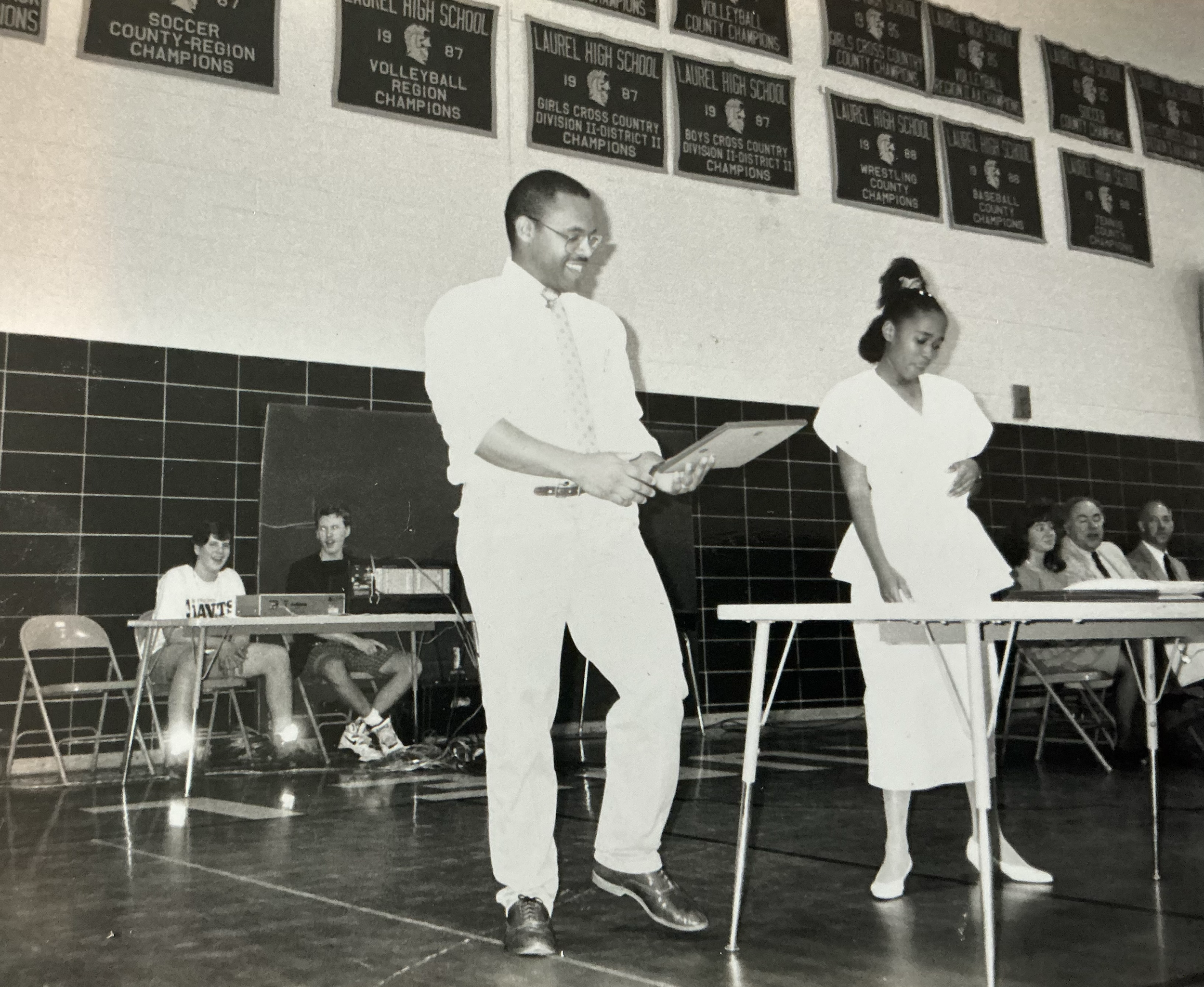 Black and white photo of students at a Laurel High School pep rally, Mark Covington is standing in the foreground, holding papers, and smiling.
