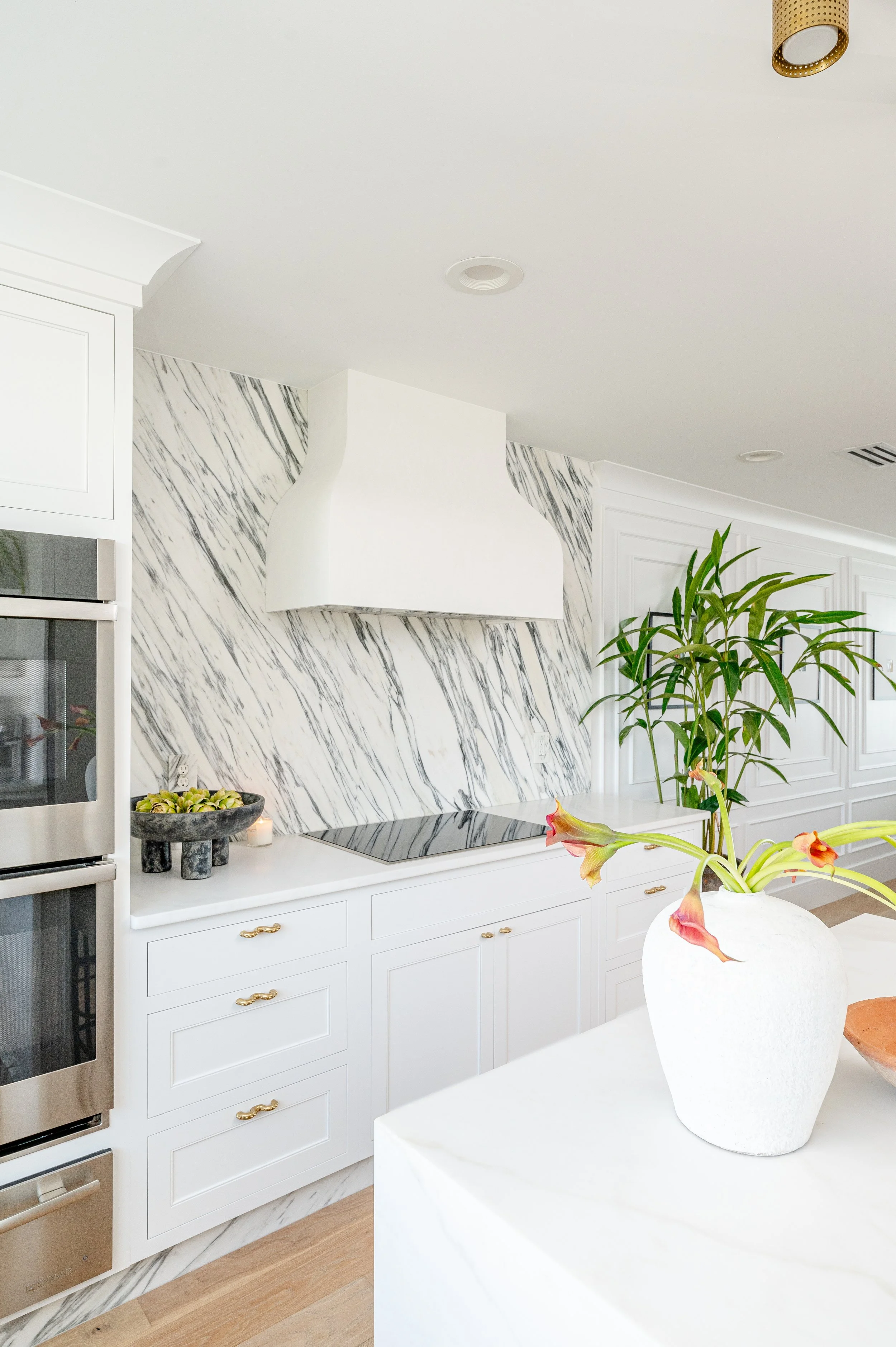 White kitchen with marble backsplash, white cabinets with gold handles, and a white range hood, with a potted plant and decorative bowl on the counter.
