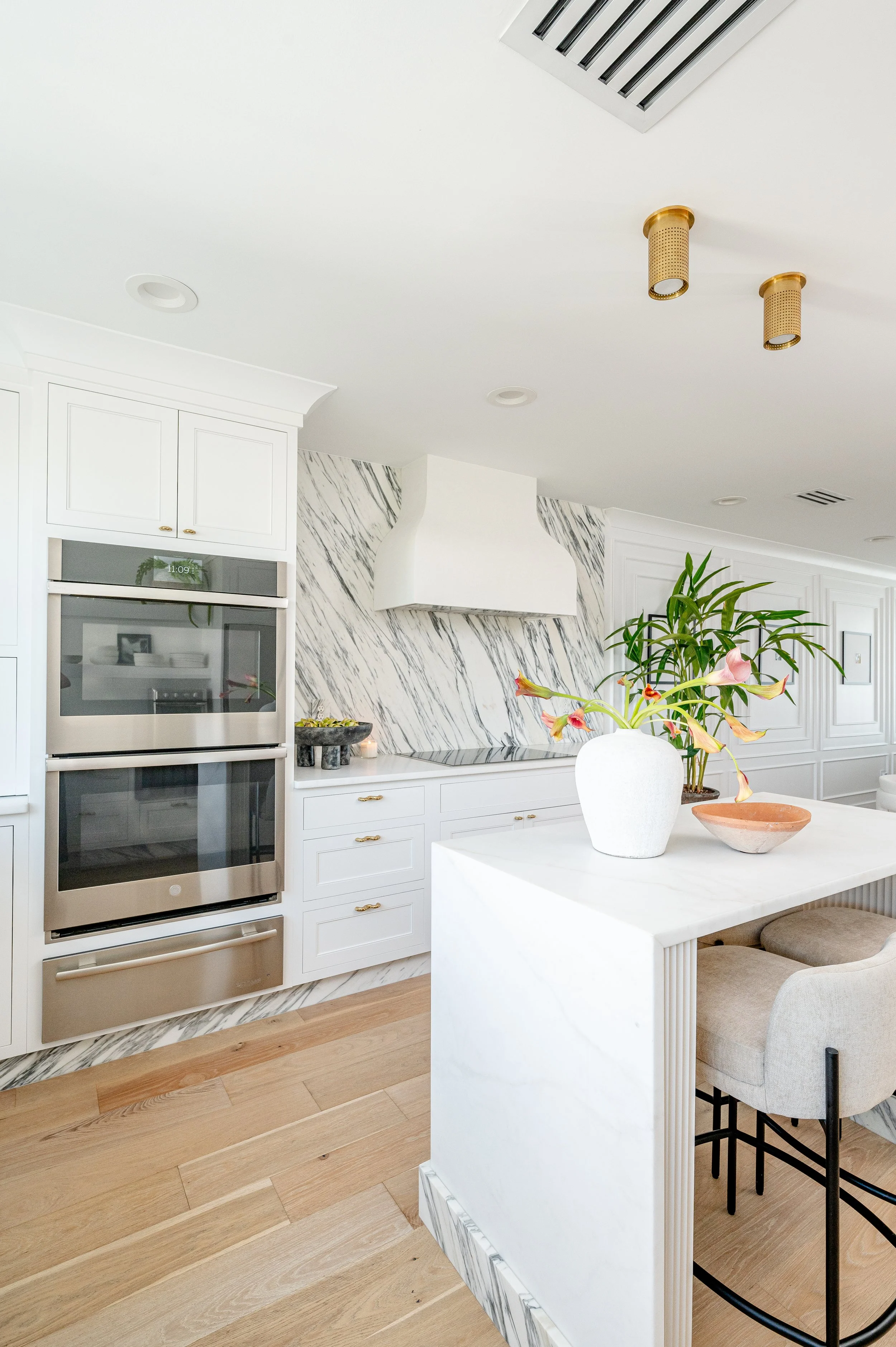 Modern kitchen with white cabinets, marble backsplash, built-in oven, and a central island with a vase of flowers.