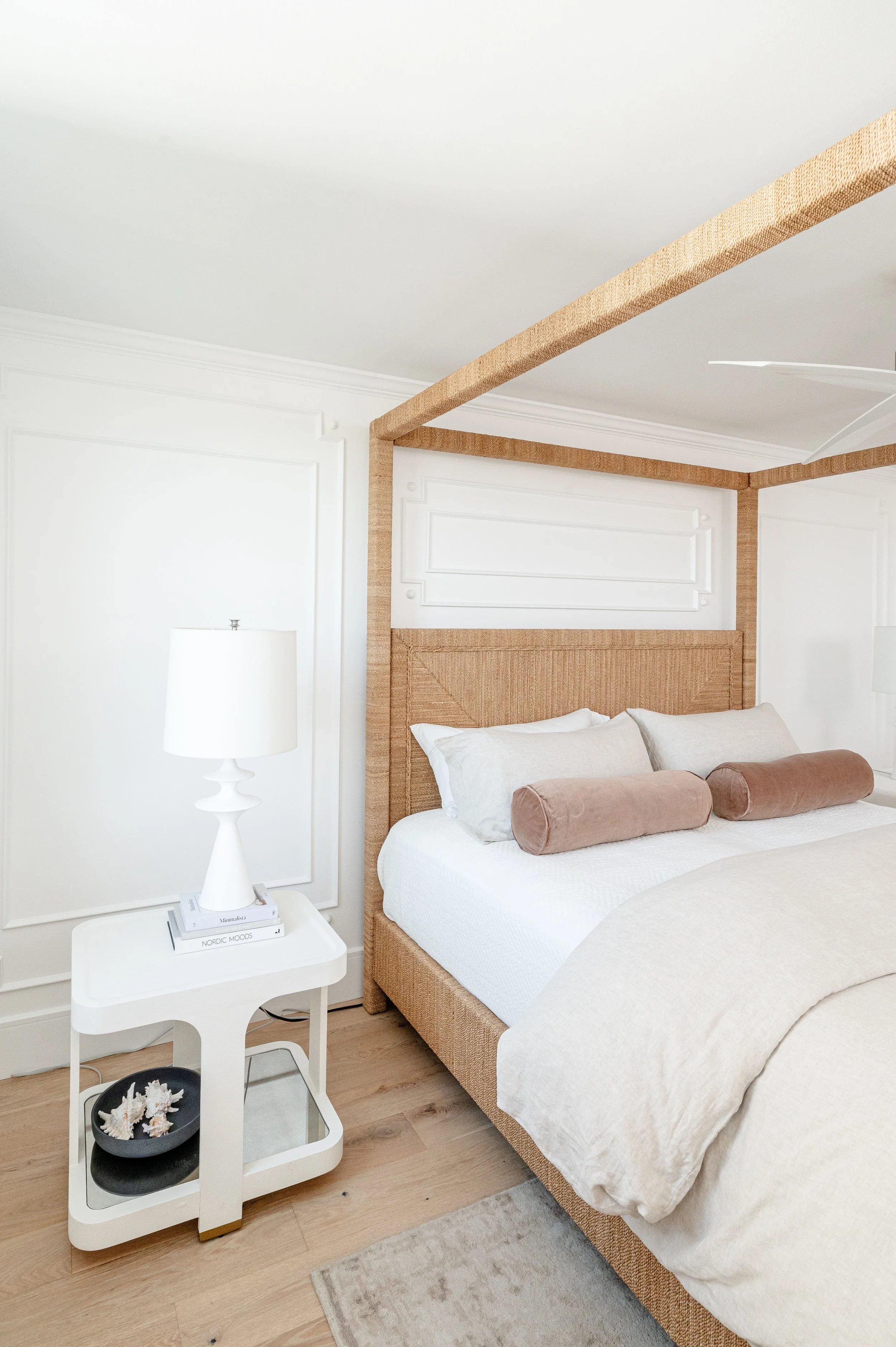 A bedroom with a bed that has a woven headboard and canopy frame, white bedding, beige cushions, a white nightstand with a lamp, books, and decorative coral, light wood floor, and white wall with decorative paneling.