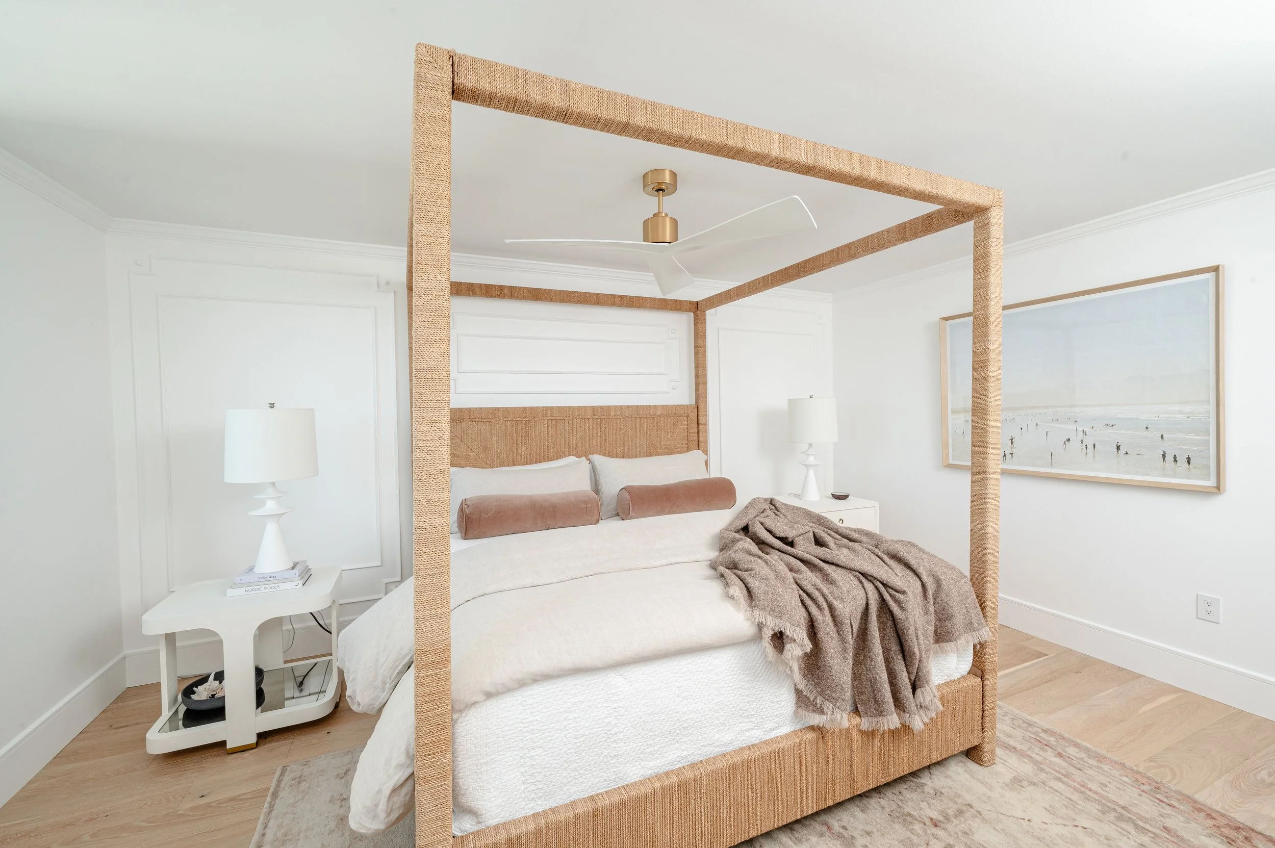 A bedroom with a four-poster bed, beige bedding, and brown pillows. Two white bedside tables with lamps, a ceiling fan, and wall art of a beach scene.