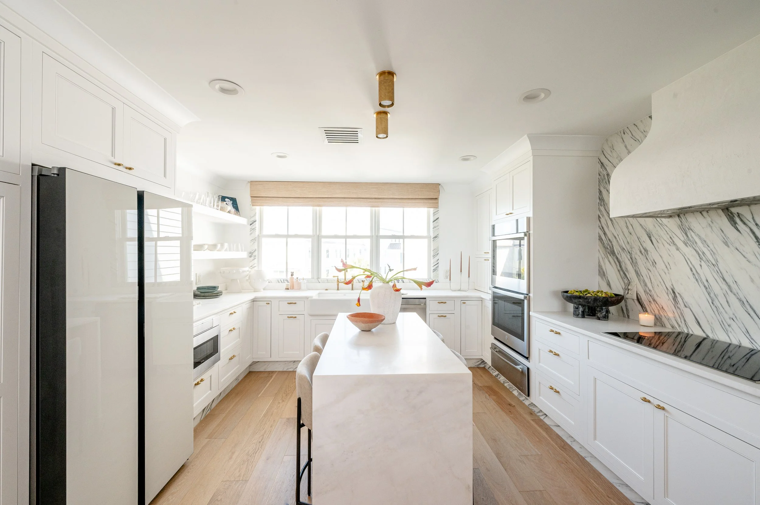 Bright white kitchen with marble accents, a large window, island with a vase and bowl, and modern appliances.