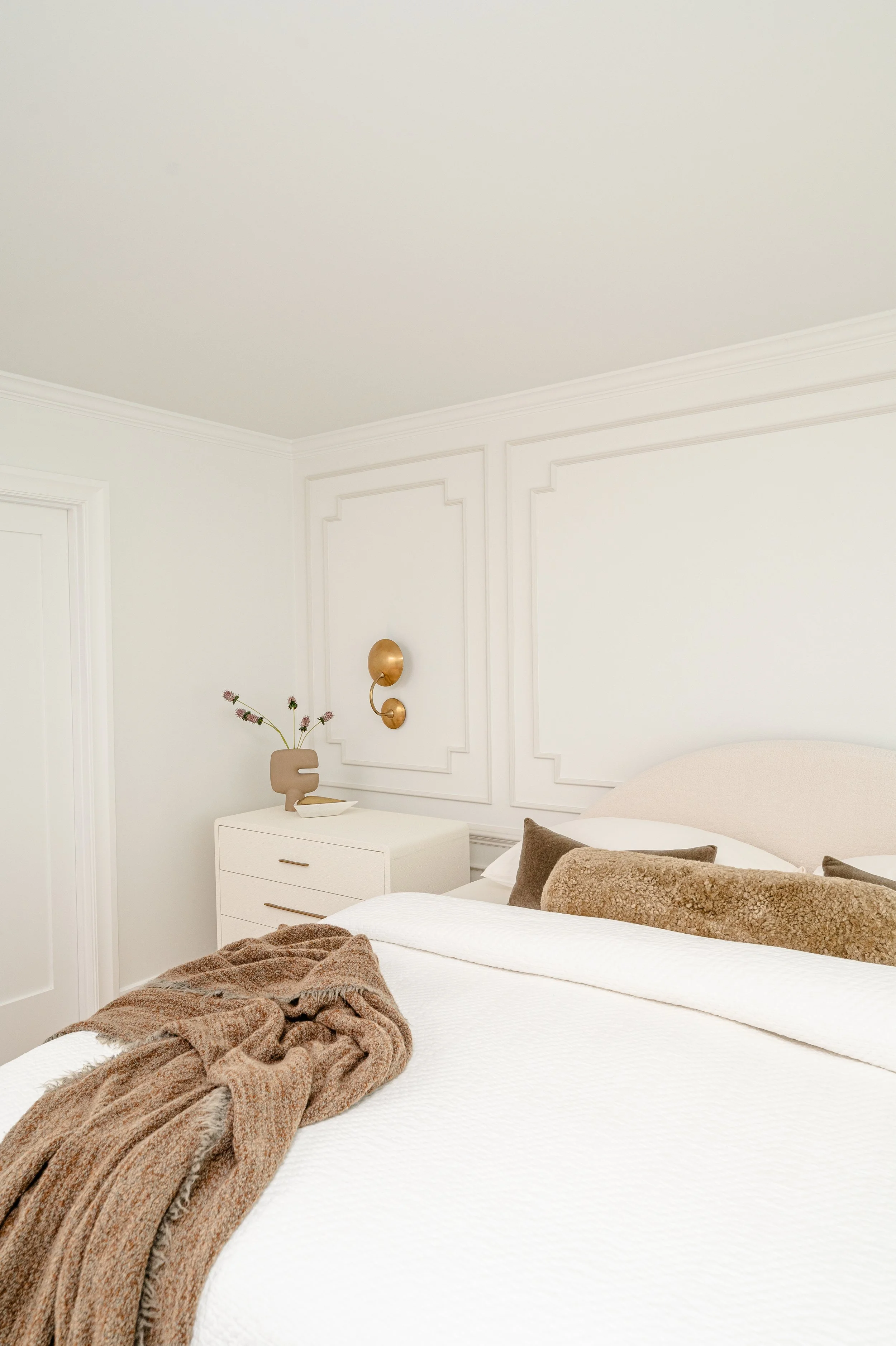 A neatly made bed with white linens and multiple brown pillows in a modern, minimalist bedroom with white walls and subtle decorative wall panels.