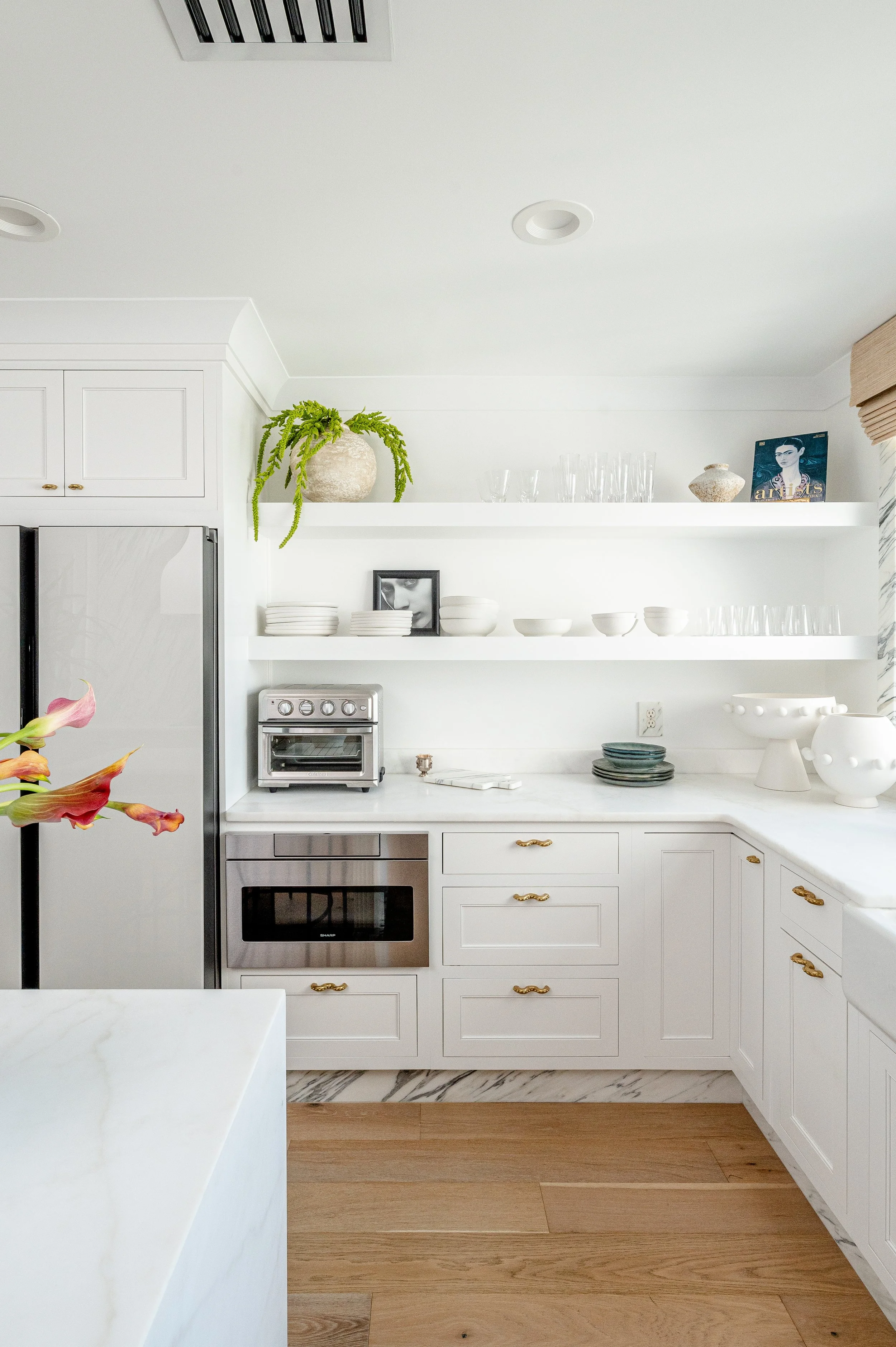 A minimalist white kitchen with open shelves displaying white dishes, glassware, and decorative objects, a stainless steel oven and toaster, and a marble countertop. There is a potted green plant on the top shelf and a small framed photo among the items.