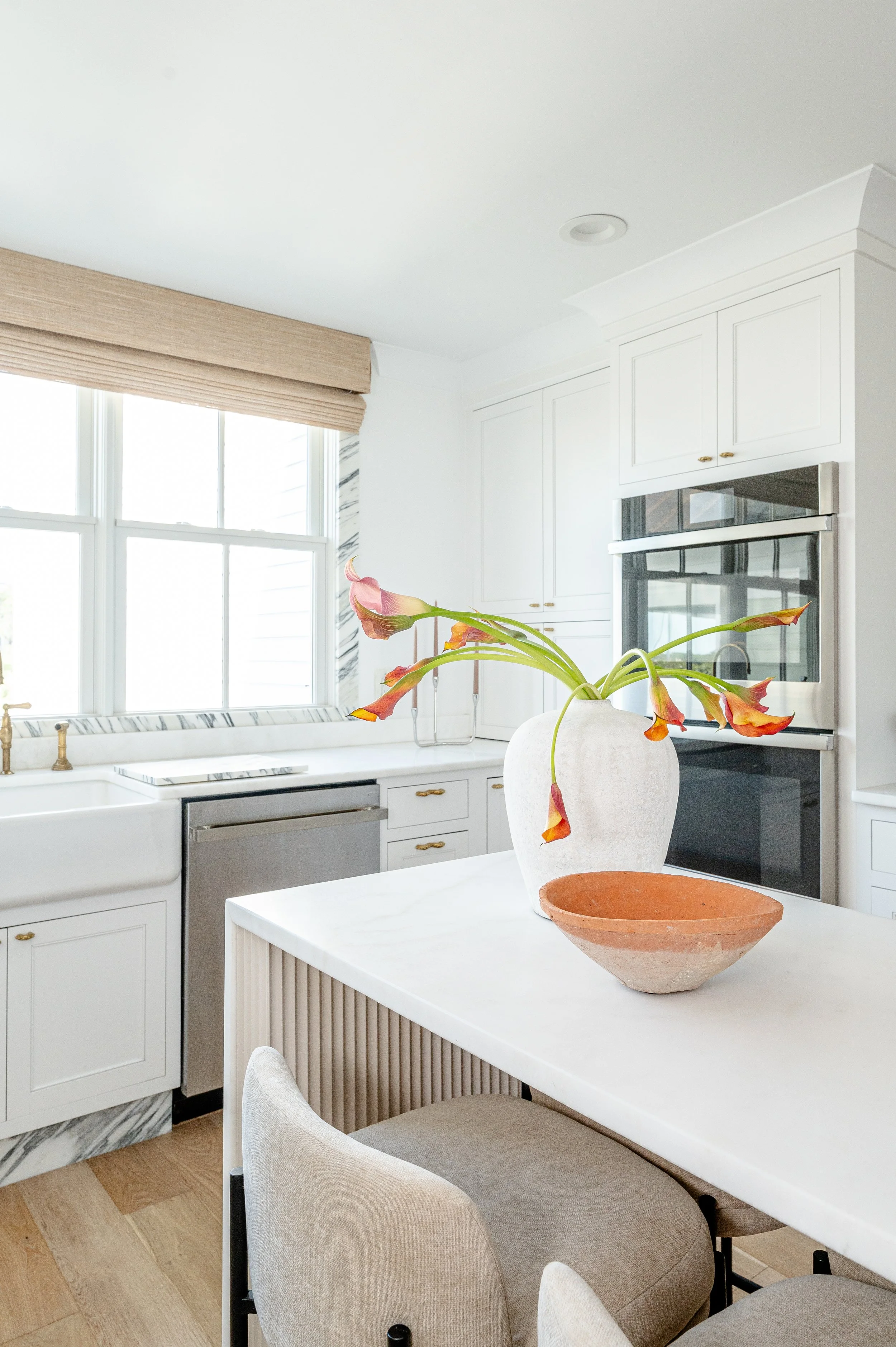 Bright kitchen with white cabinets, marble countertops, and a large window. A white vase with orange and pink flowers is on the kitchen island, along with a small terracotta bowl. There are beige upholstered chairs around the island.
