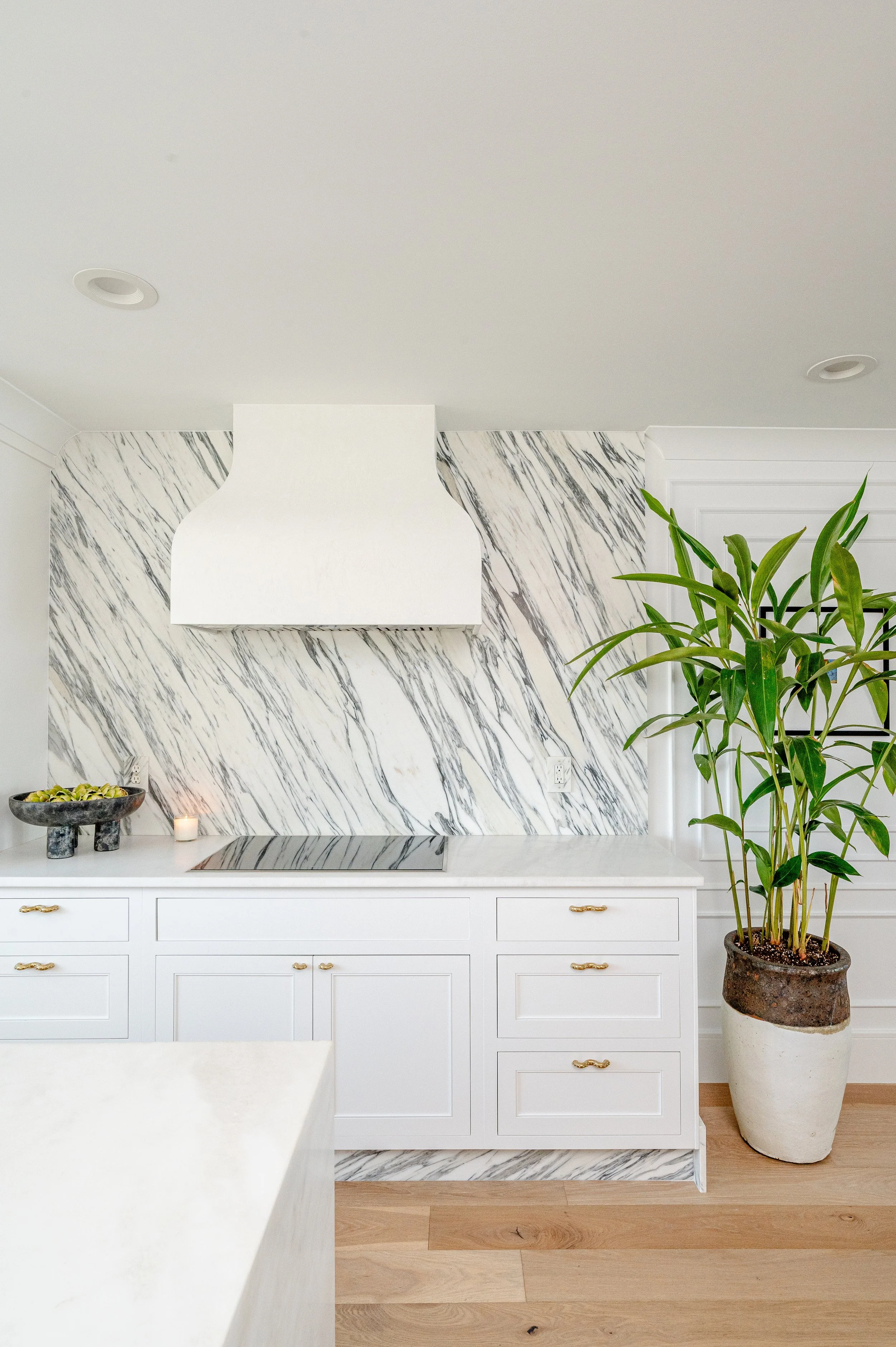 Modern kitchen with white cabinets, marble backsplash, white range hood, and a large potted plant in a rustic container.