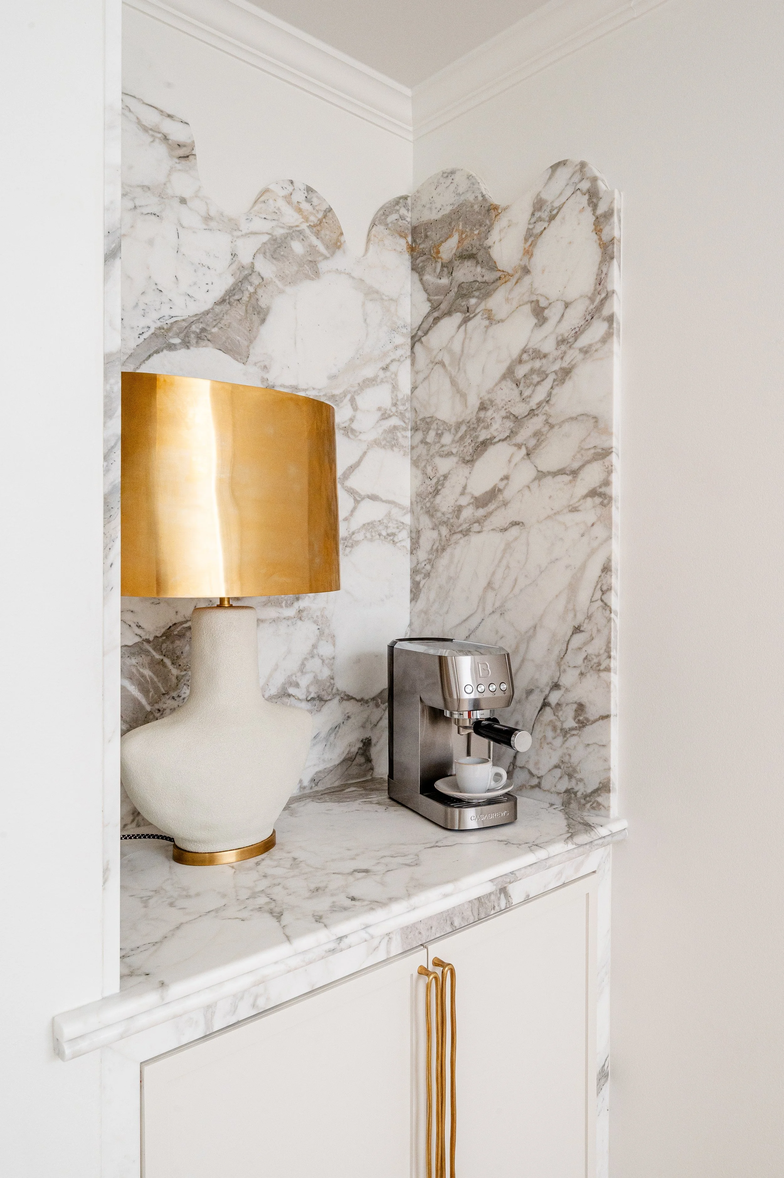A marble countertop with a gold and white lamp and a stainless steel espresso machine with a cup underneath.