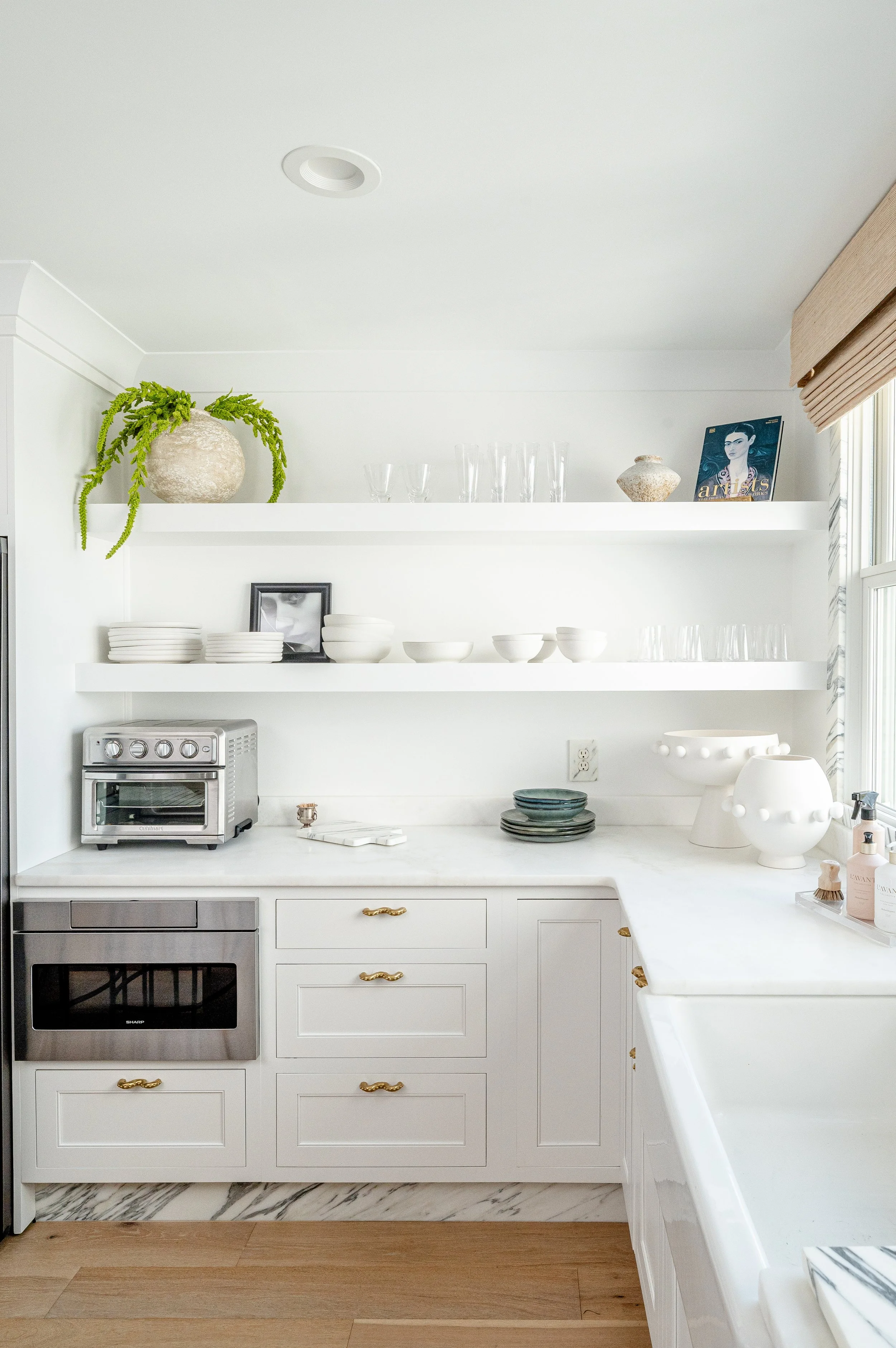 White kitchen with open shelves holding dishes and decorative items, a small oven, and a window with beige curtains.