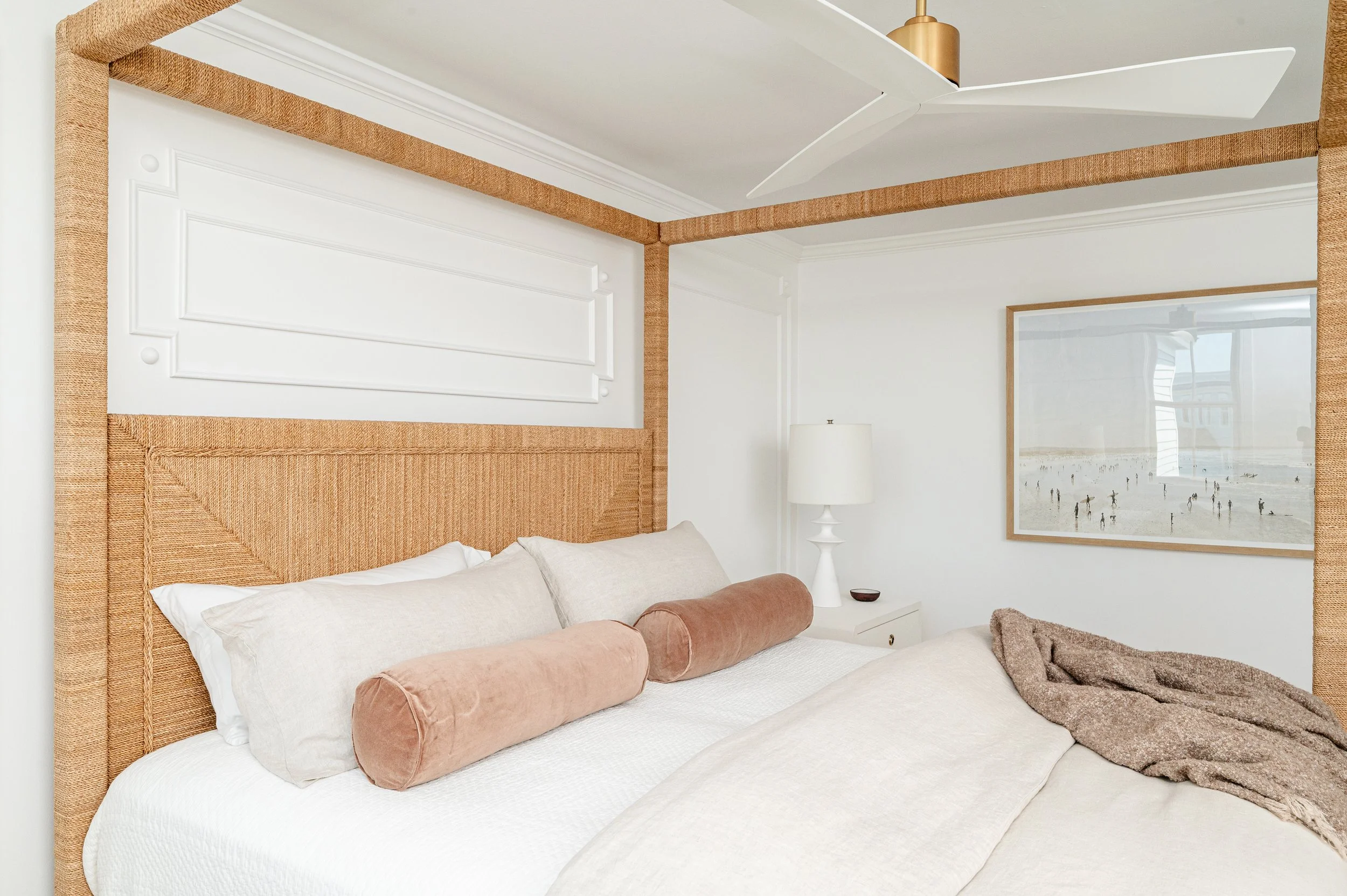 A bedroom with a bed, pillows, and a blanket. The headboard has a woven design. There is a nightstand with a lamp, a framed picture on the wall, and a ceiling fan.