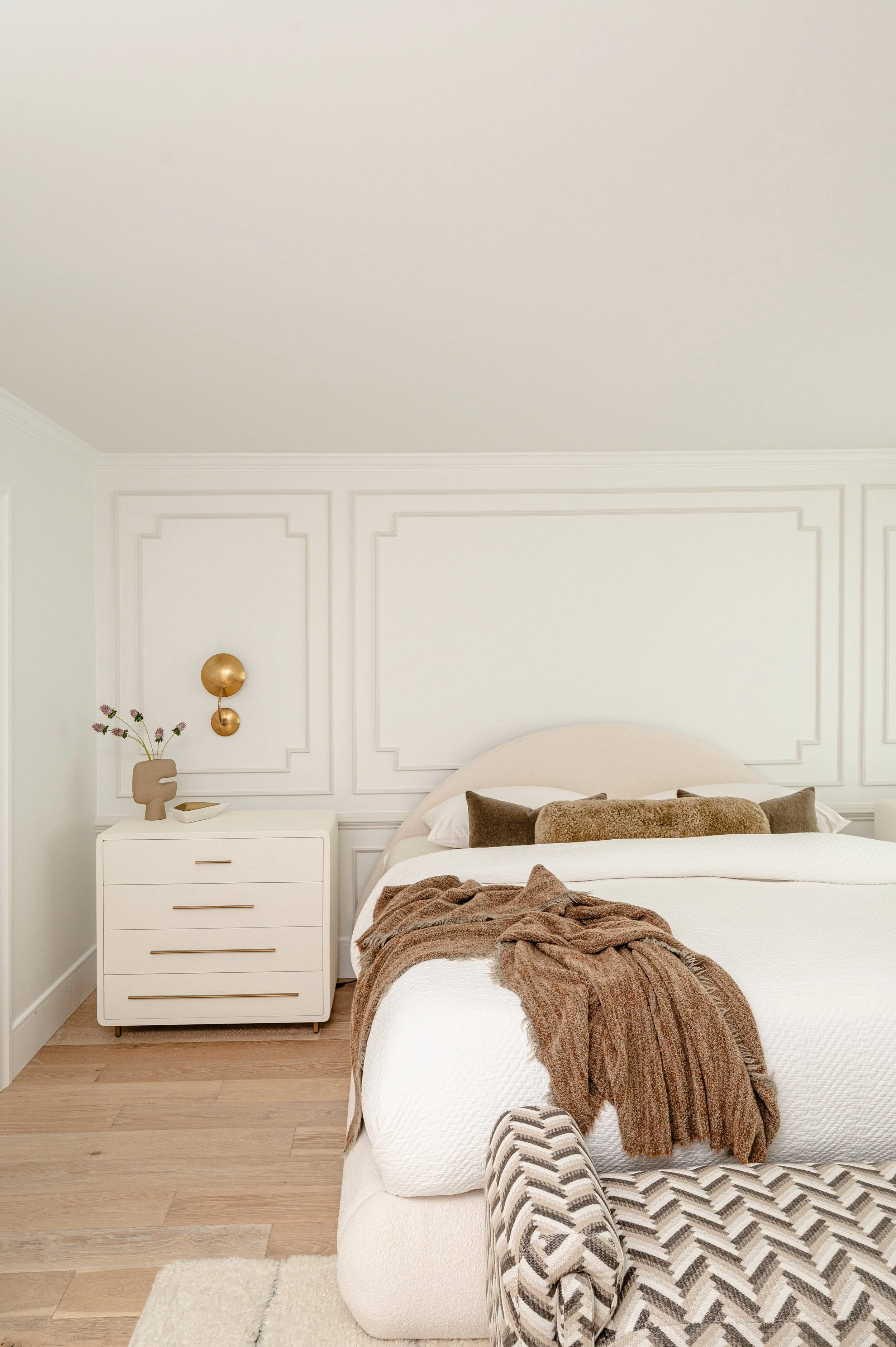 A neatly made bedroom with a white bed, brown throw blanket, brown and beige pillows, and a white nightstand with decorative items, including a gold wall sconce and a beige vase with purple flowers. The walls feature decorative molding.