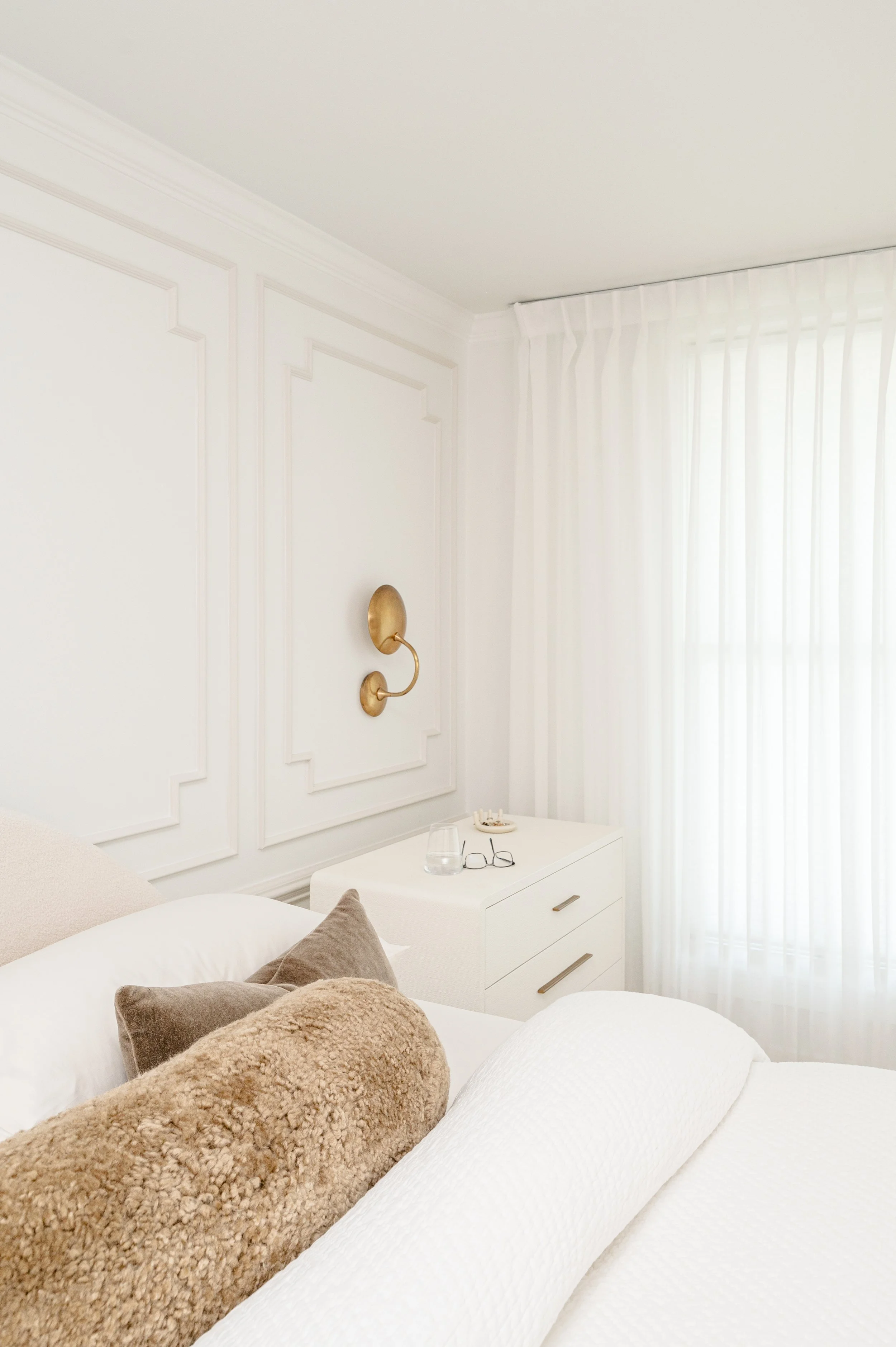 A cozy bedroom with white walls, beige and taupe pillows on a bed, sheer white curtains covering a window, a white nightstand with a glass, glasses, and a small decorative dish, and a gold wall sconce.