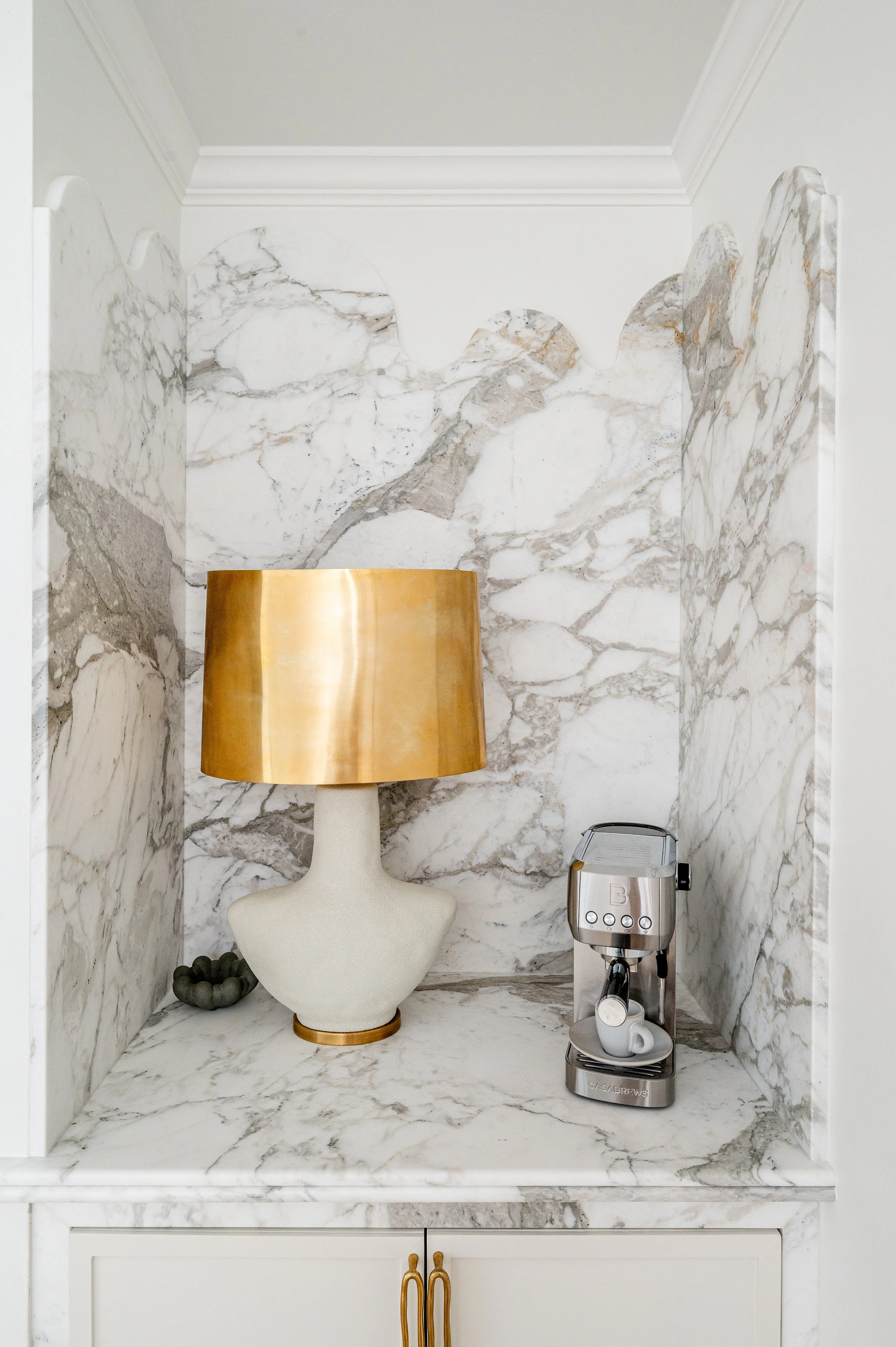 Decorative coffee station with a beige and gold vase-shaped lamp and a silver coffee machine on a marble countertop with marble backsplash.