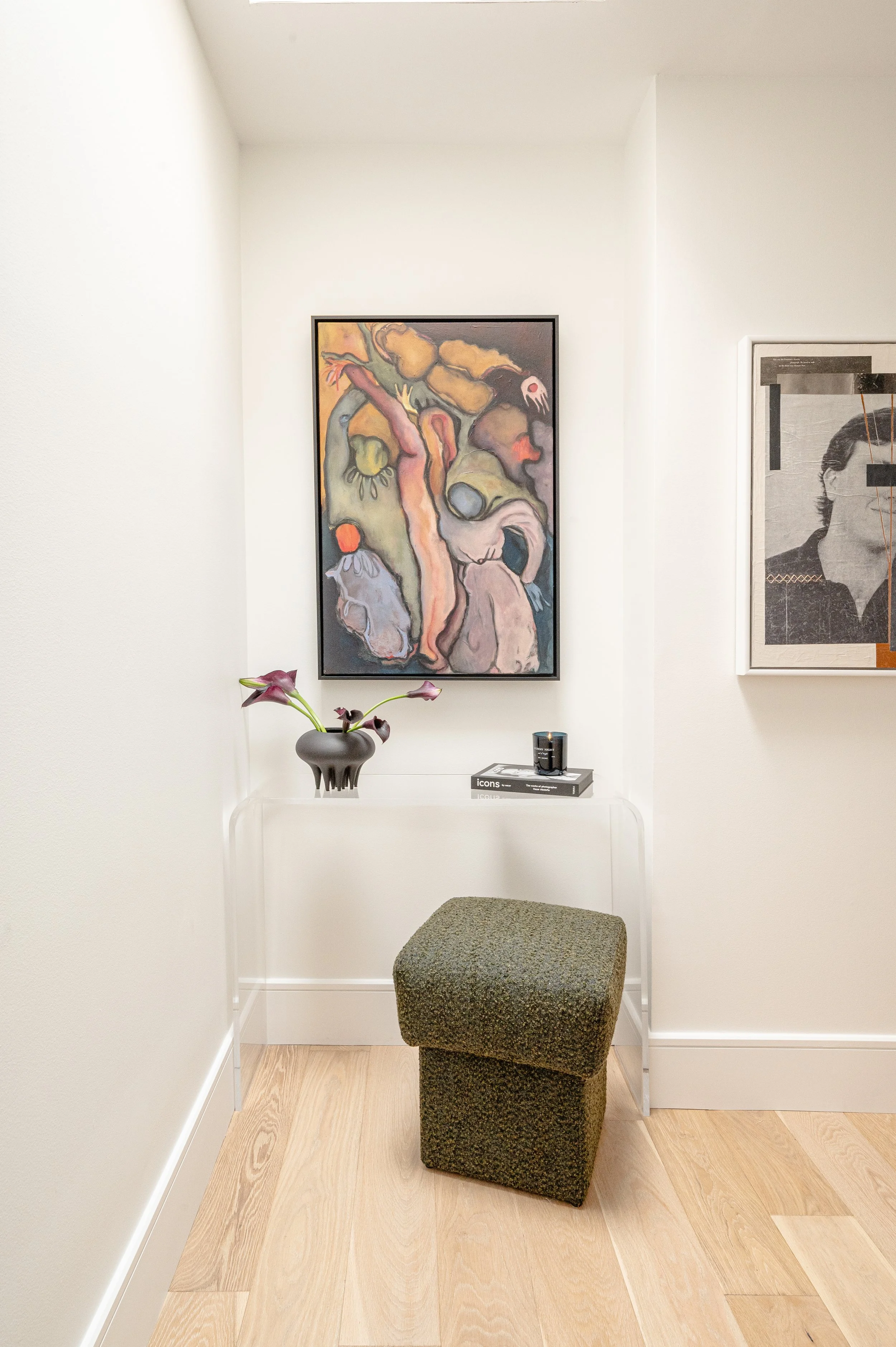 Minimalist corner with a green textured chair, a small glass table with a dark vase, purple flowers, a stack of books, and a wall with colorful abstract art.