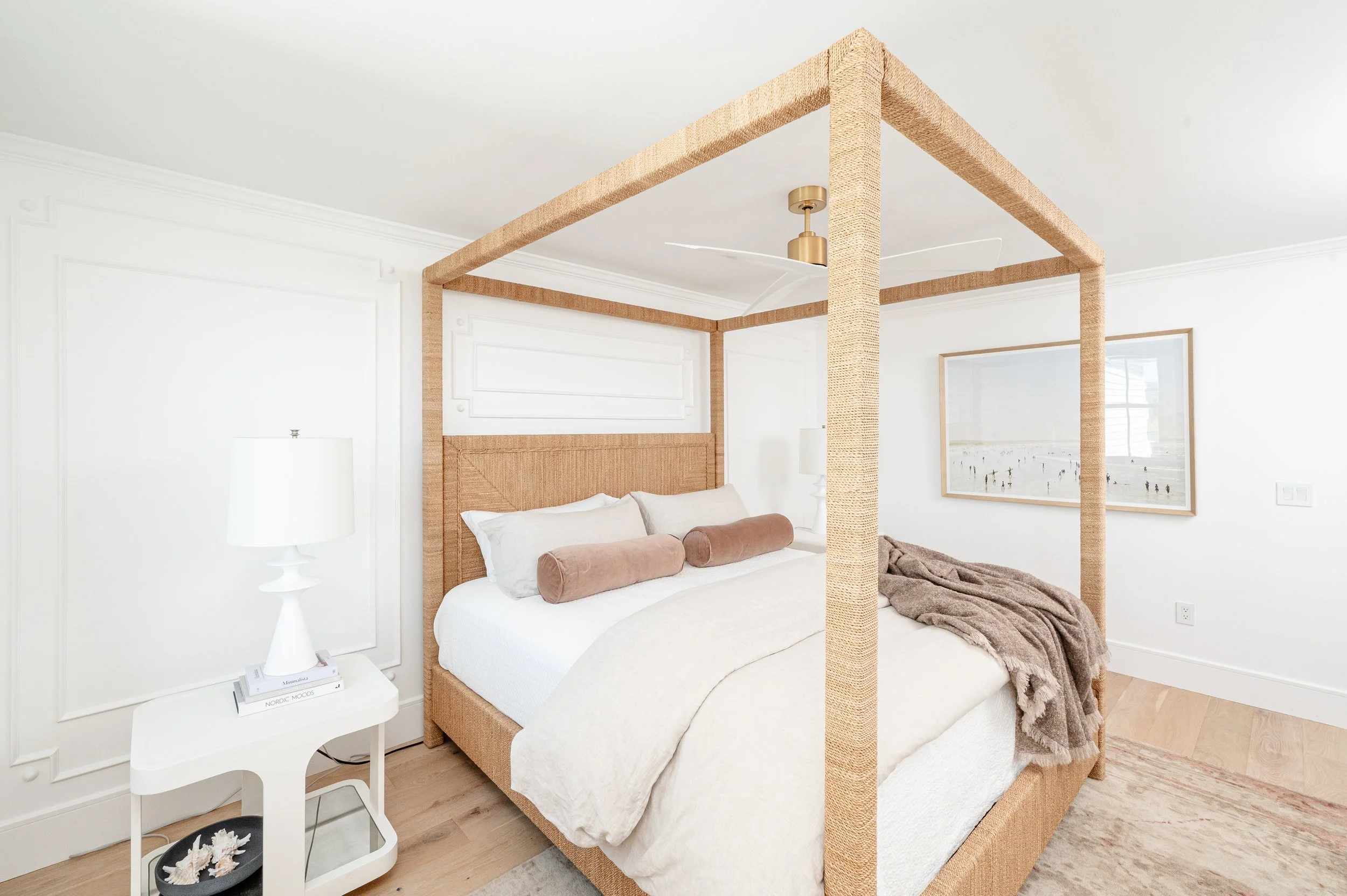 A bedroom with a four-poster bed featuring a natural woven headboard and frame, white bedding, beige bolster pillows, a beige throw blanket, a white bedside table with a lamp, and a framed seaside photograph on the wall.