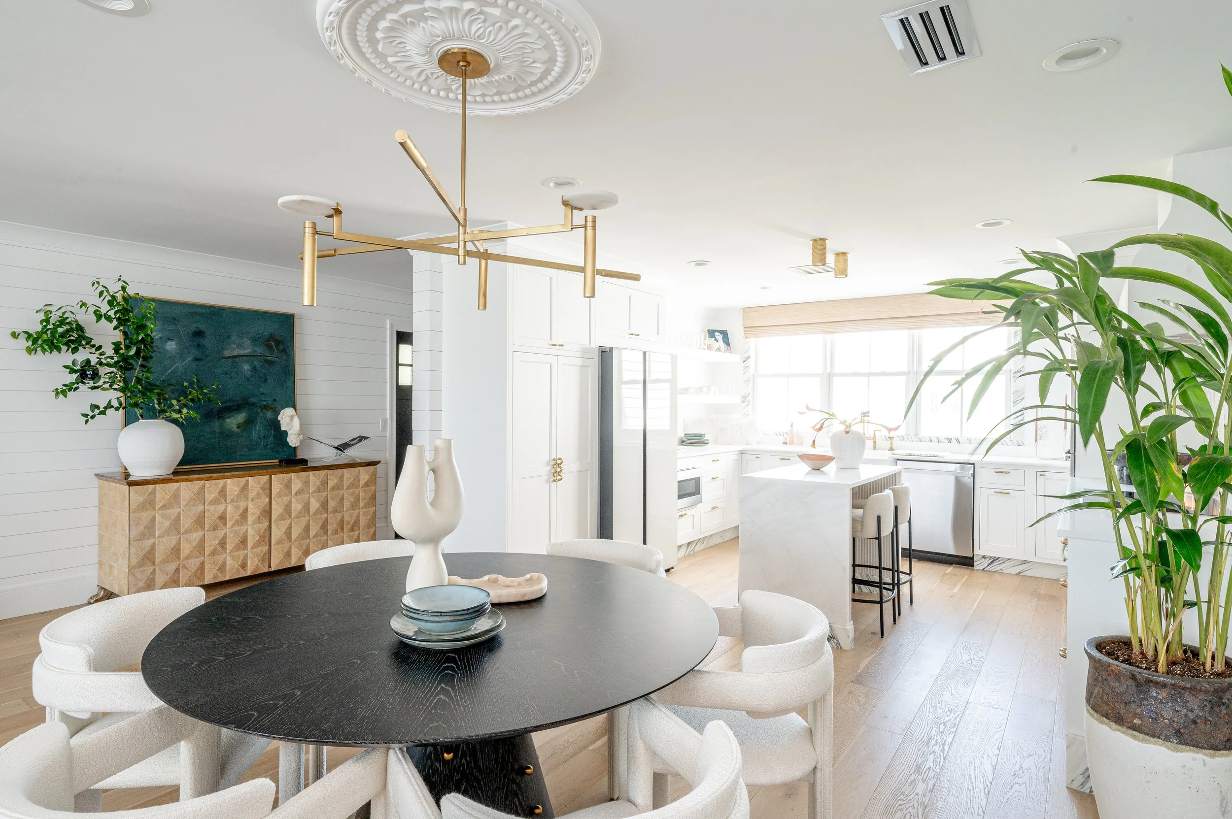 Bright, modern kitchen and dining area with white cabinetry, marble countertops, large window, and green plants. Dining table with white chairs and decorative vases, stylish light fixture overhead.