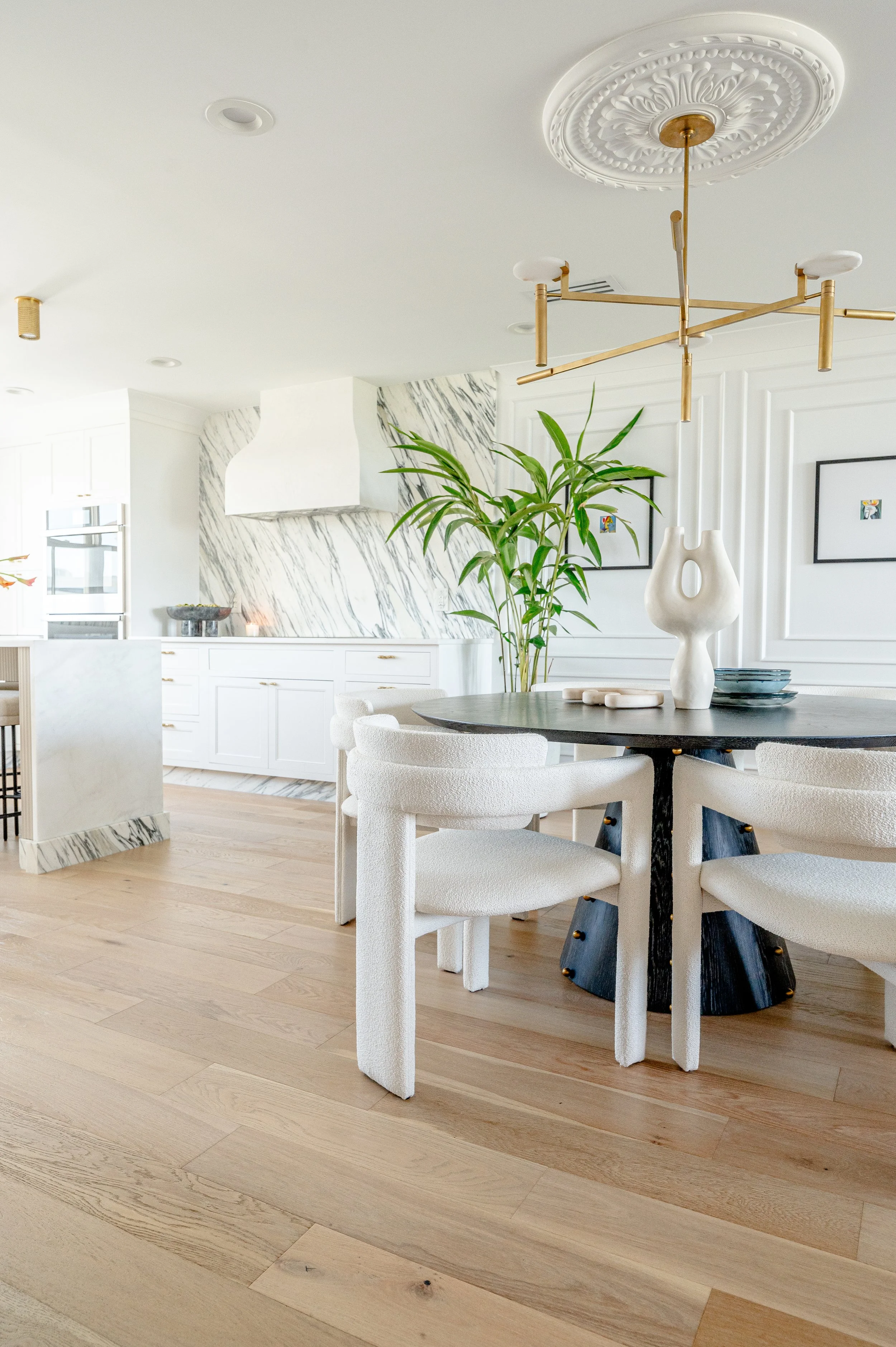 Modern dining room with a round black table, white upholstered chairs, a large potted plant, and a gold and white chandelier, with a white kitchen in the background.