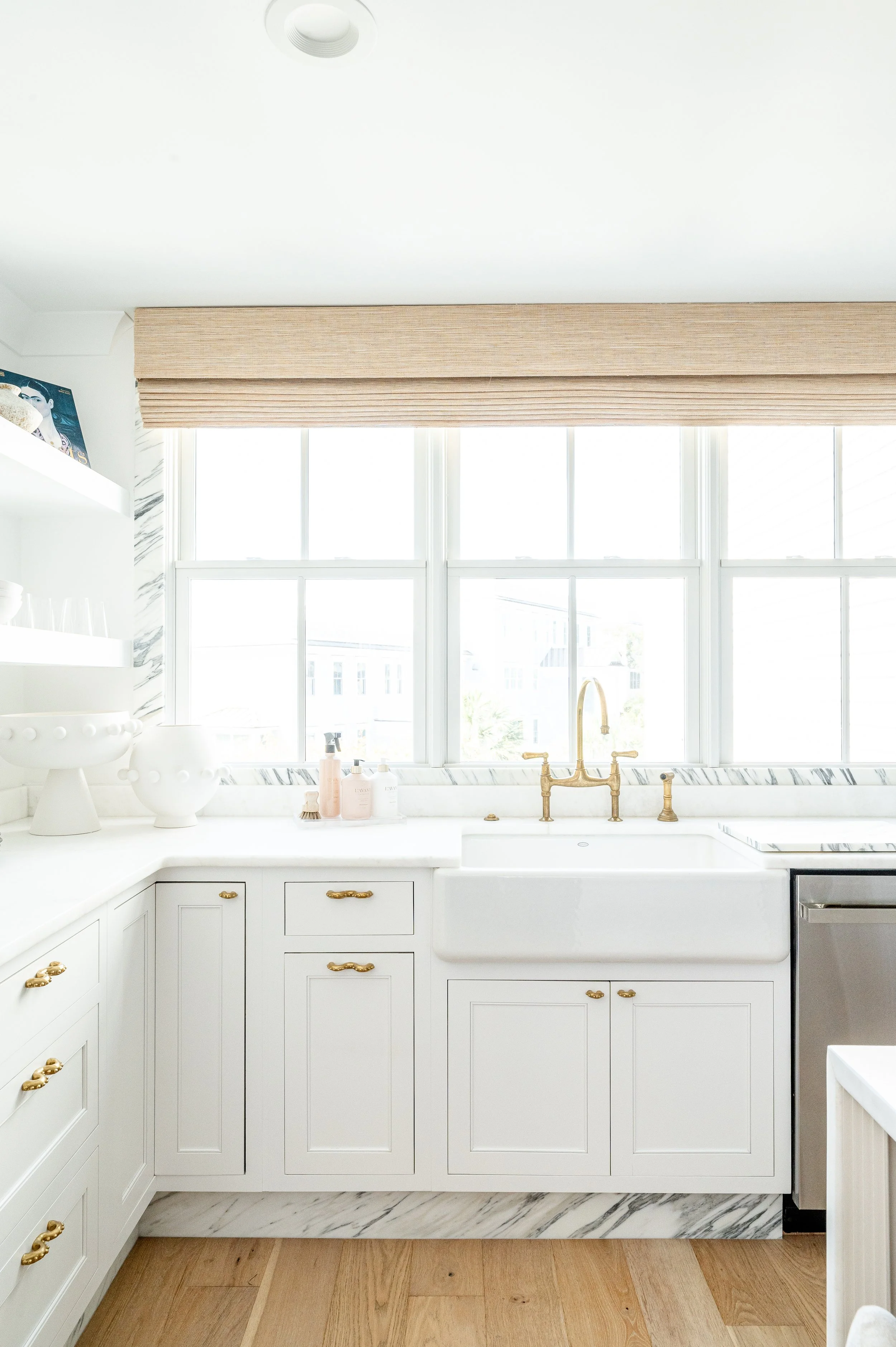 Bright kitchen with white cabinets, marble countertops, gold hardware, a farmhouse sink, a large window with beige Roman shade, and wooden flooring.