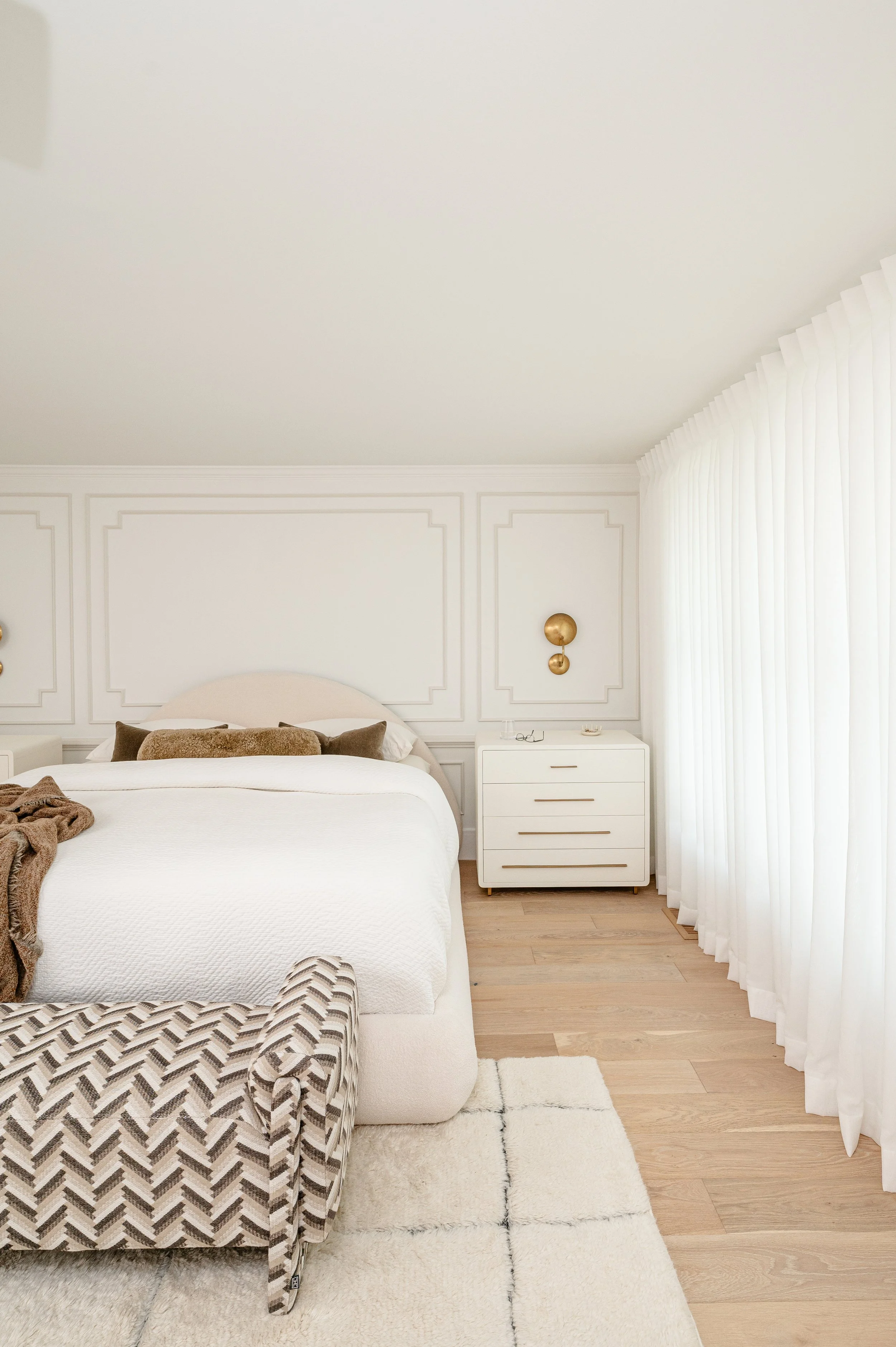 A bedroom with a large bed, white bedding, and decorative pillows. There is a patterned armchair at the foot of the bed, a white rug, and a nightstand with gold wall sconces on a paneled white wall. Large curtains cover a window on the right.
