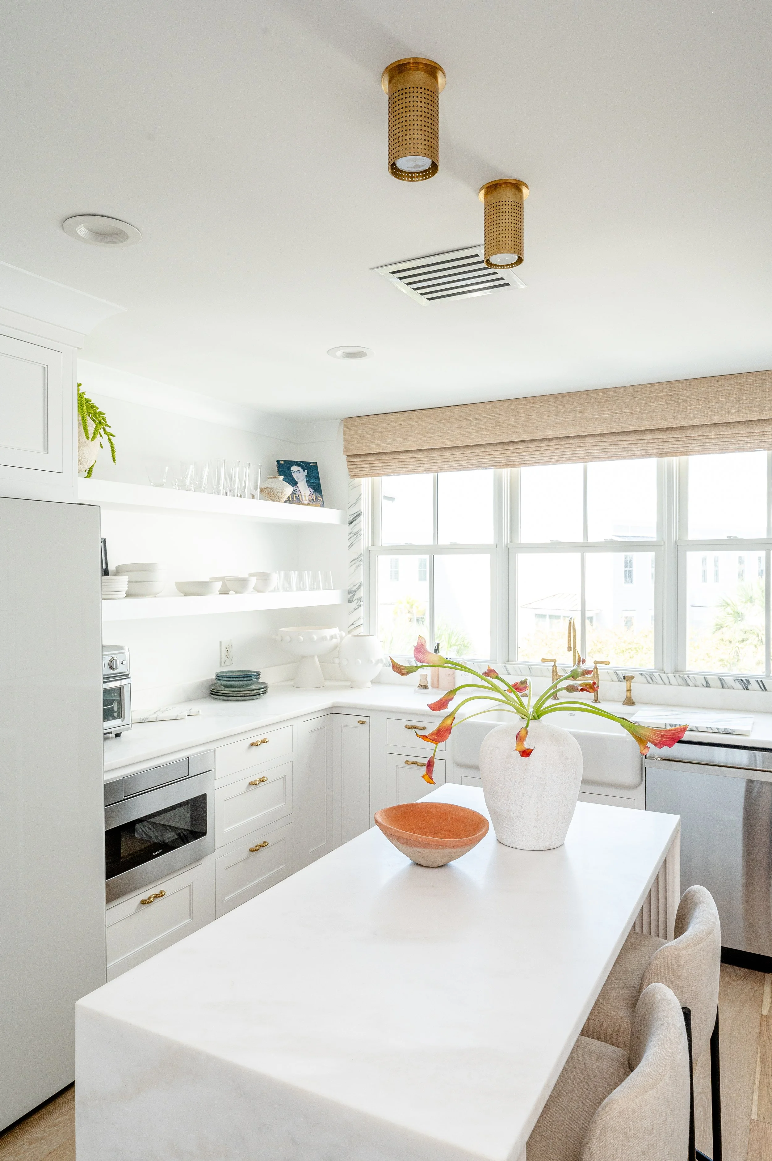 Bright kitchen with white cabinets, large window, beige chairs, a white island with a vase of flowers, and gold light fixtures.