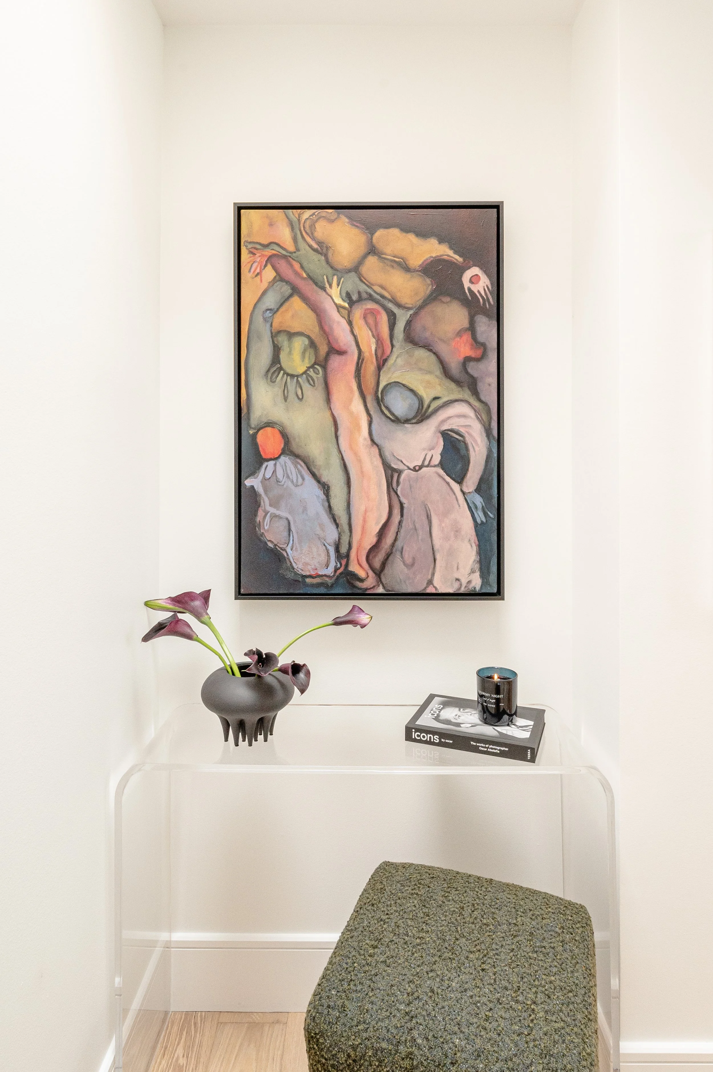 An art gallery corner featuring a colorful abstract painting on a white wall, a round vase with dark purple calla lilies on a clear table, a black candle, and a book titled "Icons". A textured gray stool is in front of the table.