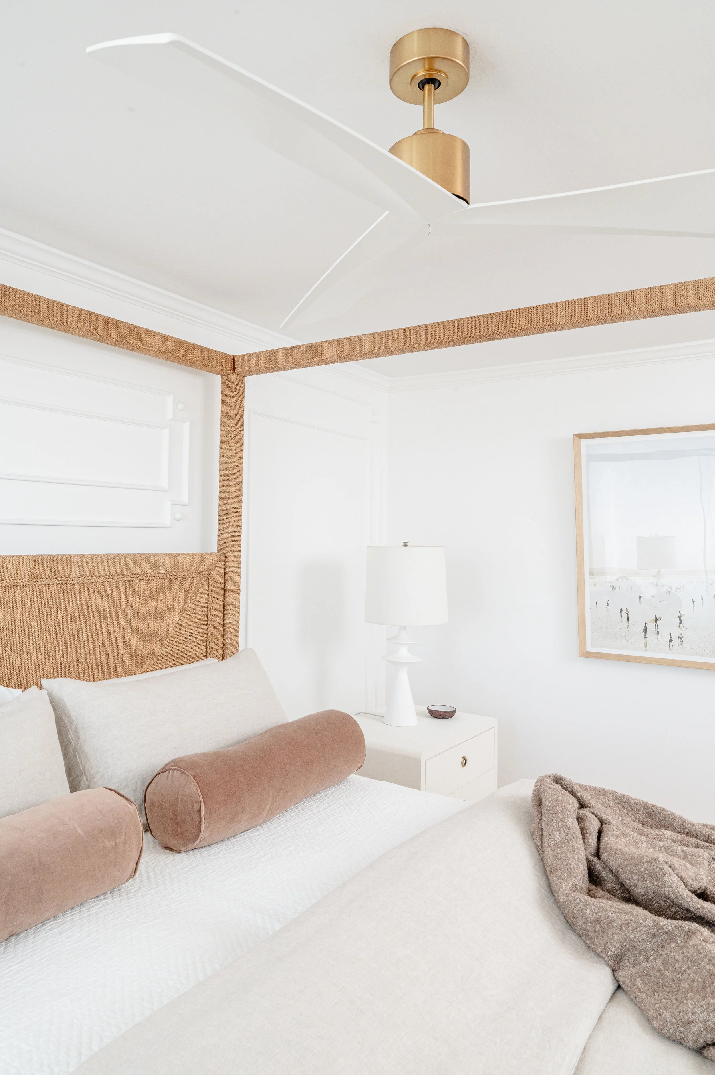 A bedroom with a white bed, beige pillows, a white side table with a lamp, a framed picture on the wall, and a ceiling fan with a gold finish.