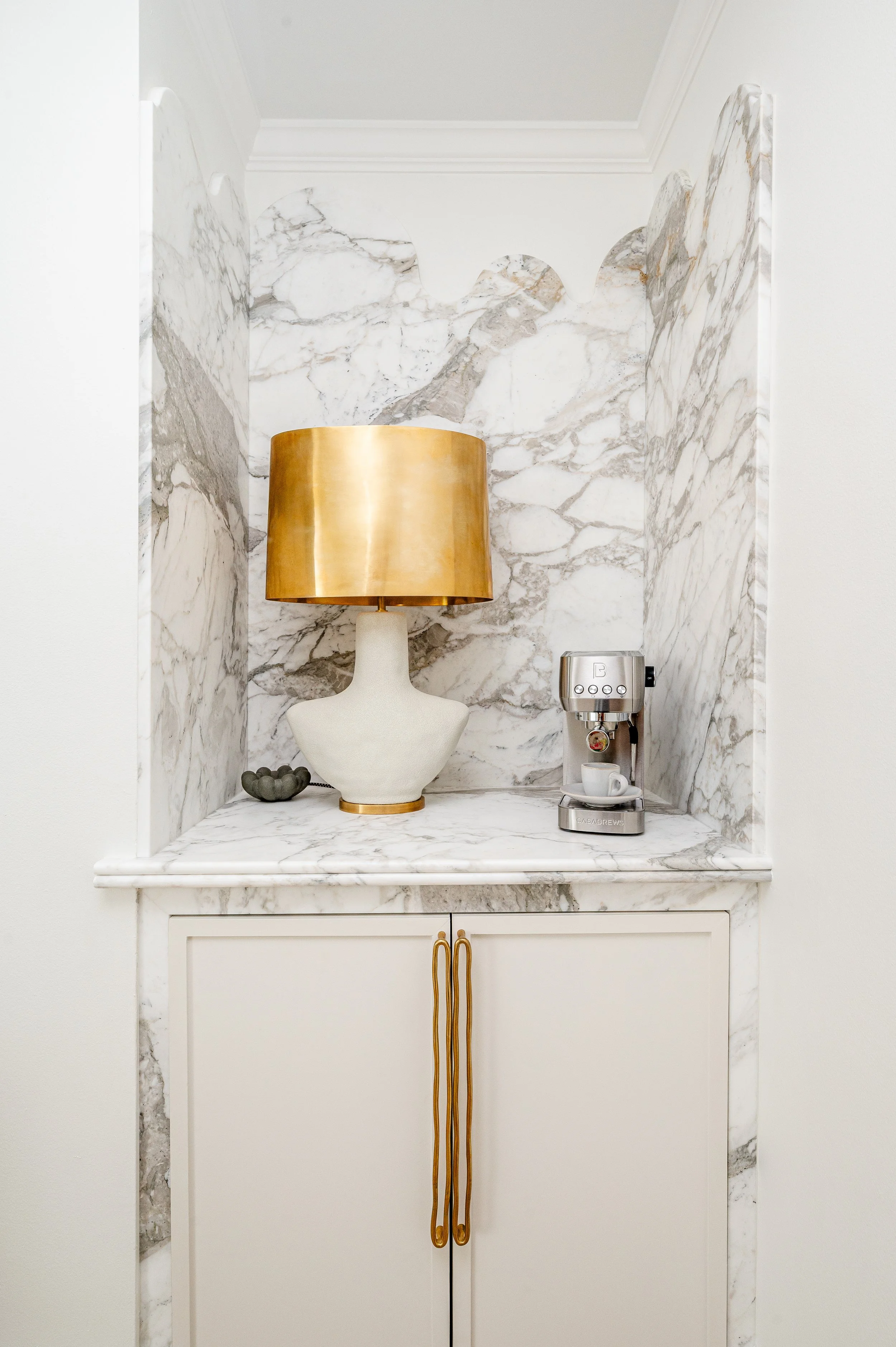 A marble countertop and backsplash with a white cabinet below. On the countertop, there is a large white and gold table lamp, a small green decorative object, and a silver espresso machine with a cup of coffee.