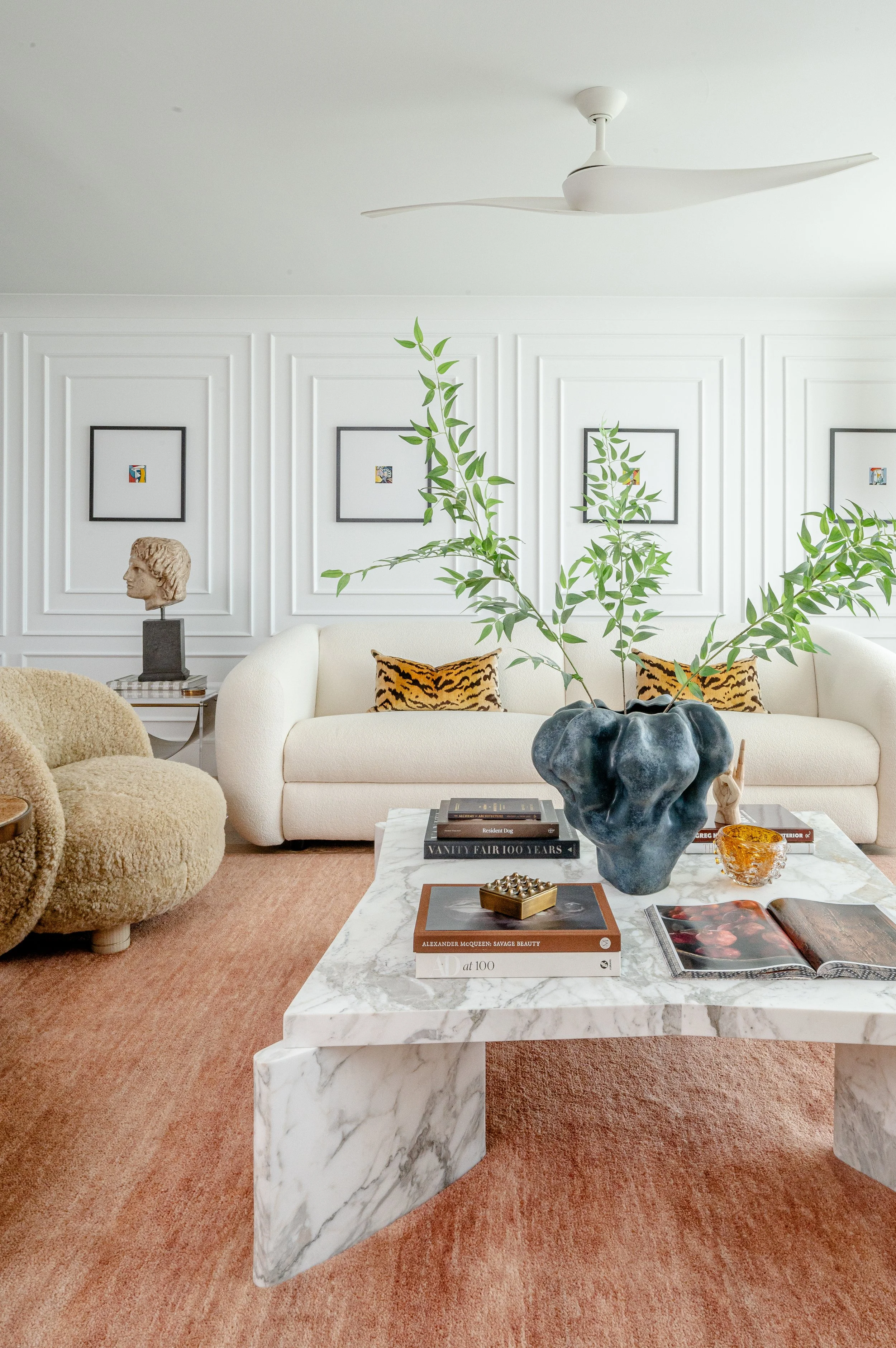 Living room with white sofa, animal print pillows, marble coffee table, green plant in a dark ceramic vase, beige textured armchair, decorative objects, and framed artwork on the white paneled walls.