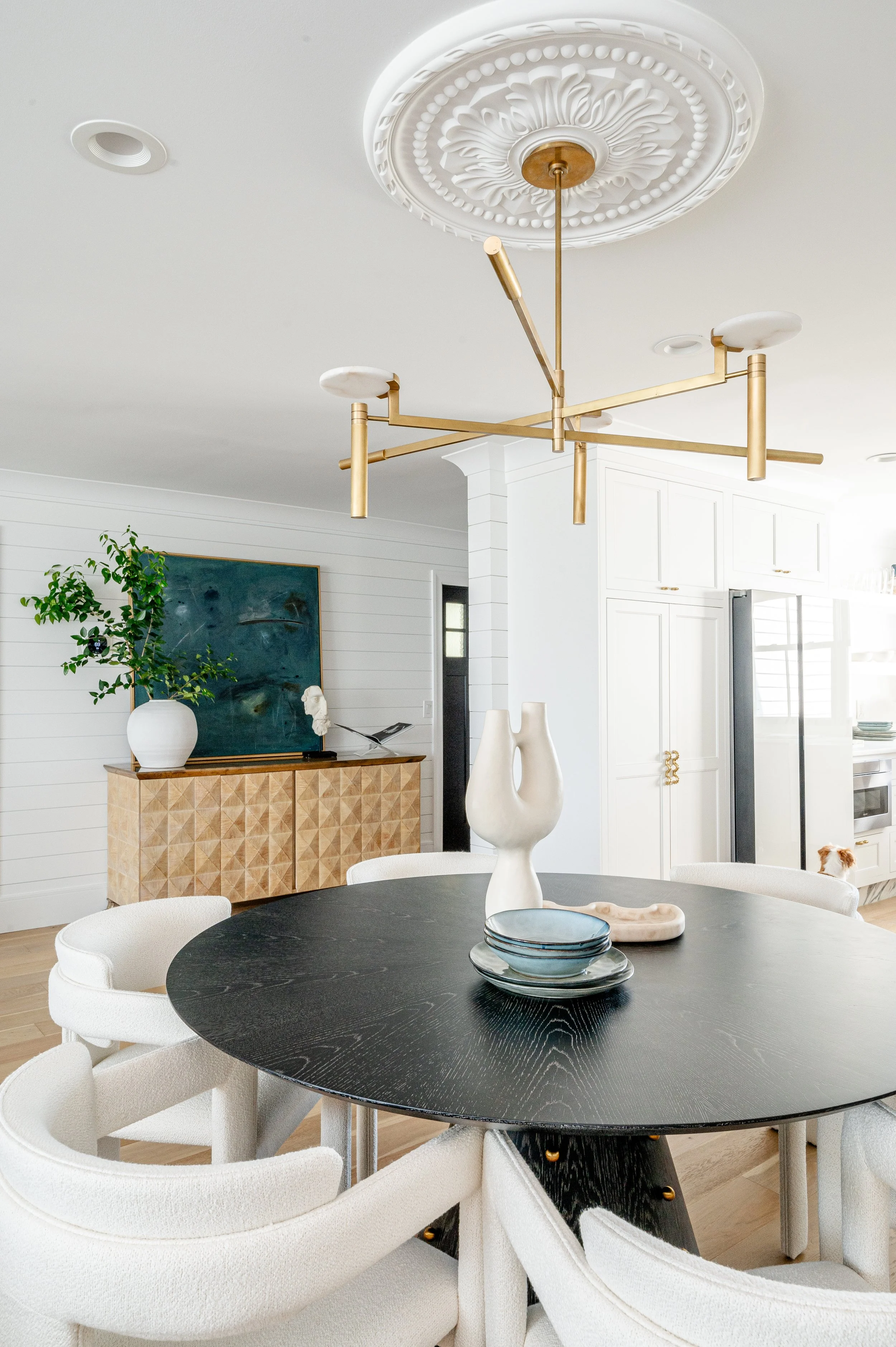 Modern dining room with black oval table, white chairs, a decorative chandelier, and a sideboard with artwork, vases, and houseplant.