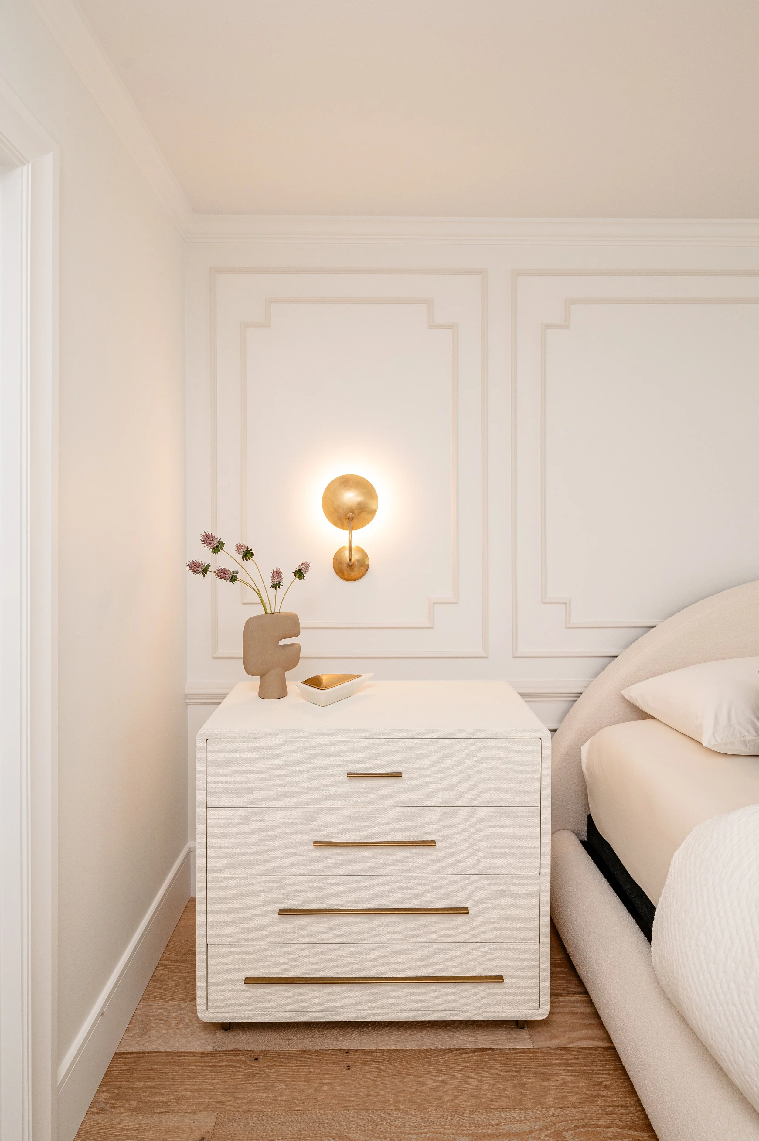 A white bedside table with four drawers and gold handles next to a bed with white bedding. On top of the table is a beige decorative vase with pink flowers and a small white dish. Behind the table, a modern gold wall sconce is mounted on a white wall