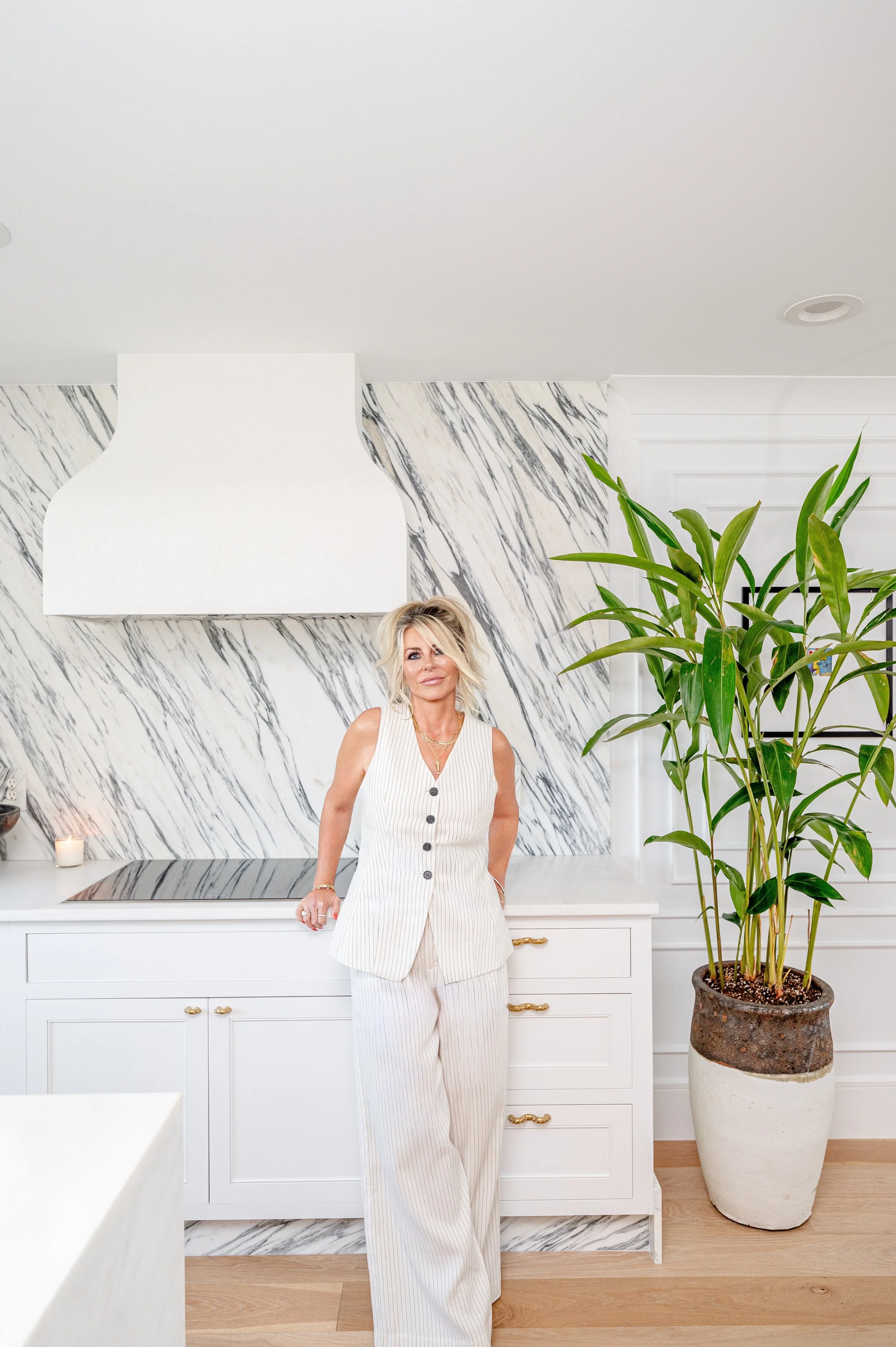 A woman standing in a modern kitchen with white cabinetry, a marble backsplash, a large potted plant, and a white range hood.