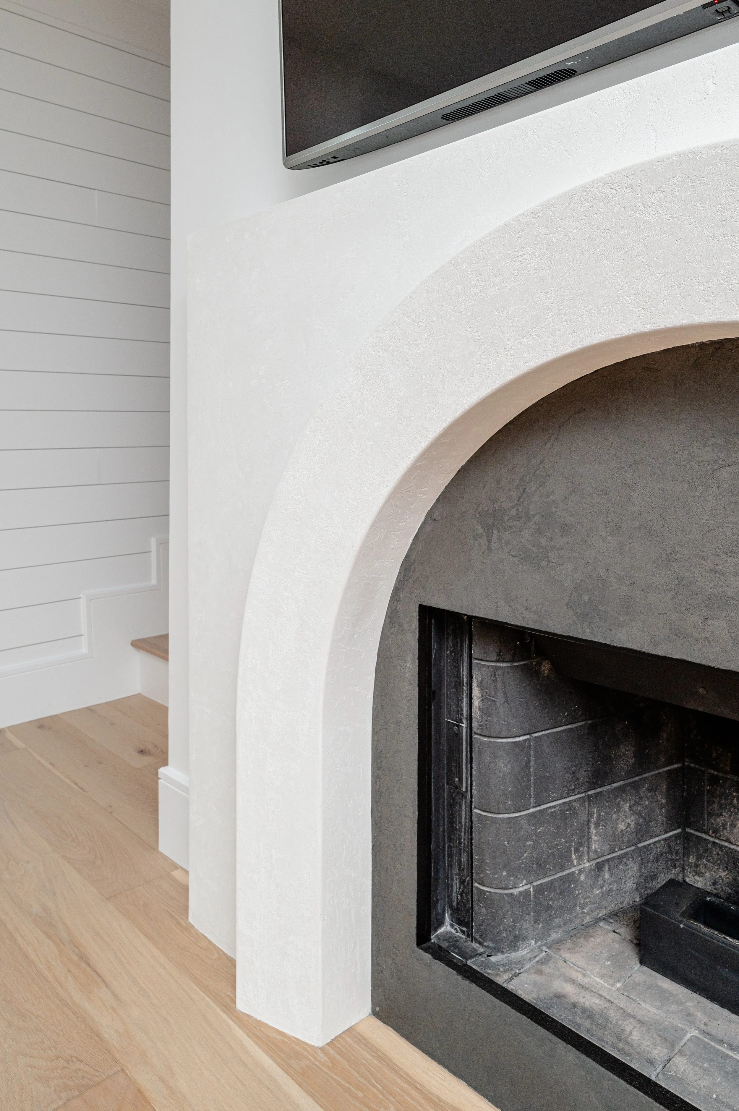 Close-up of a modern fireplace with a black interior and a white textured arch surround, situated in a room with light wooden flooring and white paneled walls.