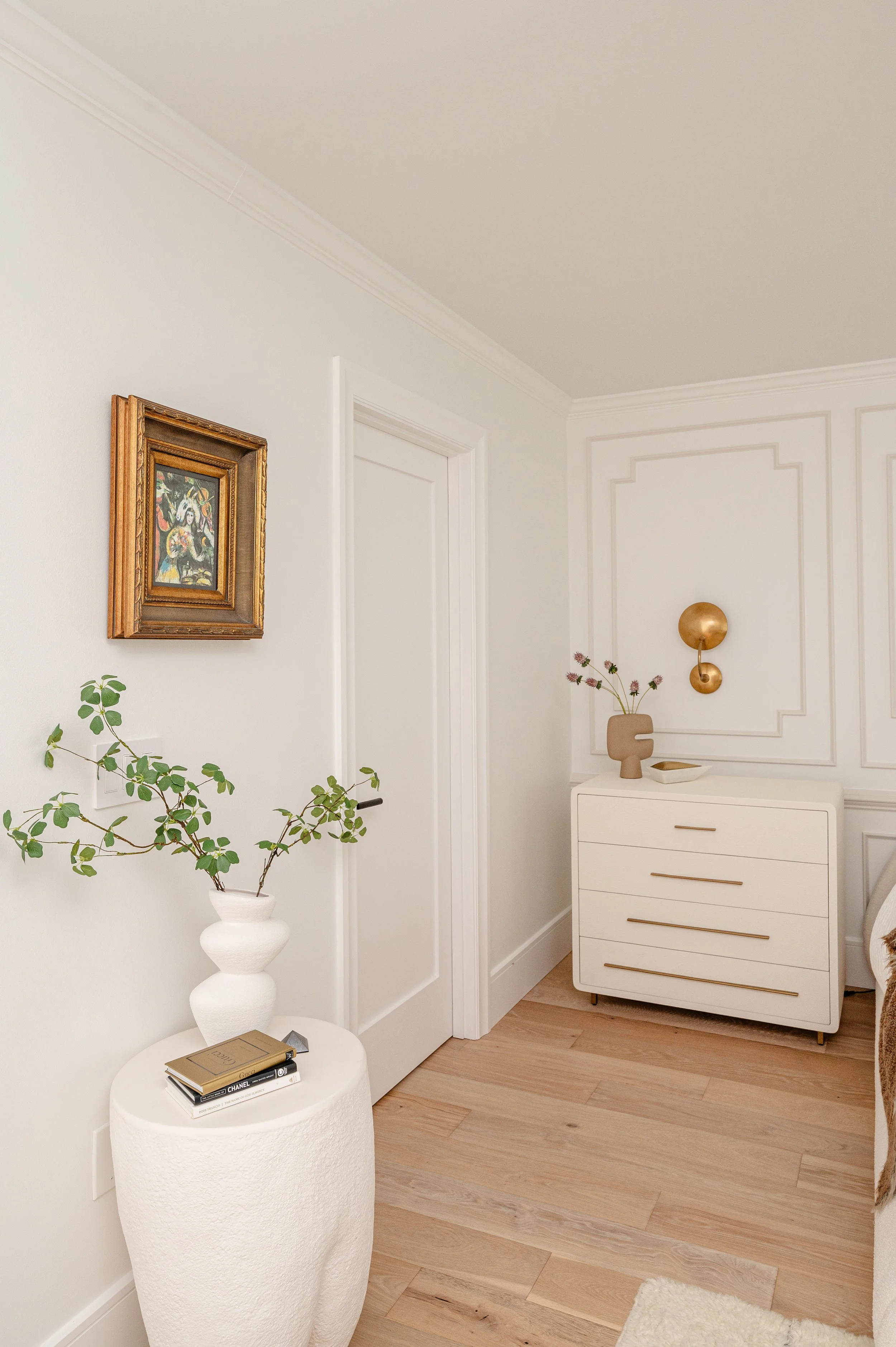 A corner of a modern, minimalist bedroom with white walls and light wooden flooring. There is a small white textured round side table holding two books and a small decorative object. A white vase with green leafy branches sits on the table. On the wa