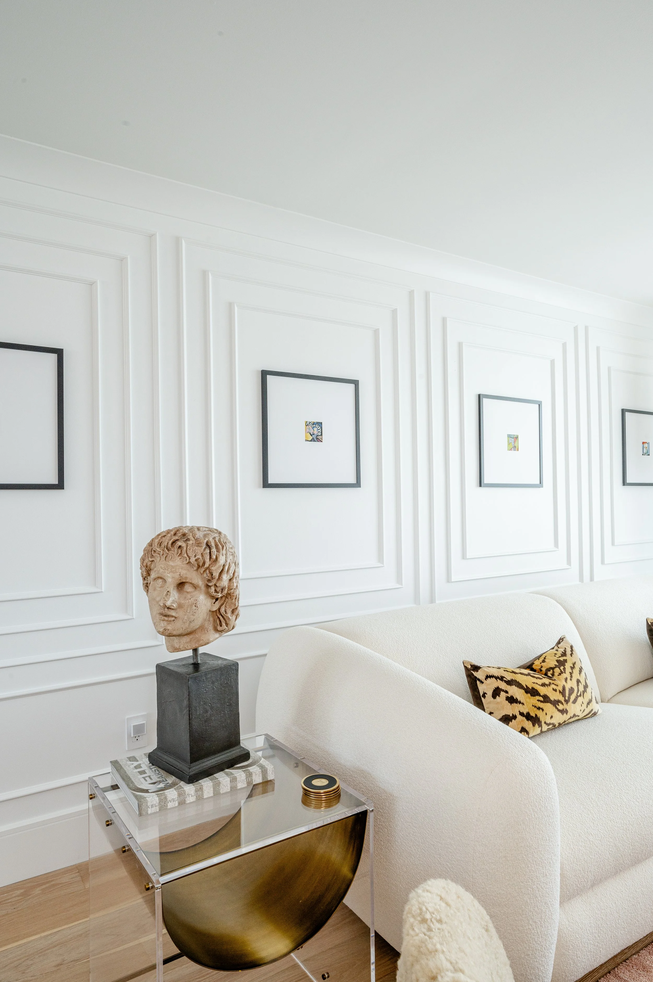 Modern living room with white paneled walls, abstract framed artwork, a white curved sofa with leopard print pillow, a glass coffee table with decorative items, and a classical bust sculpture on a pedestal.