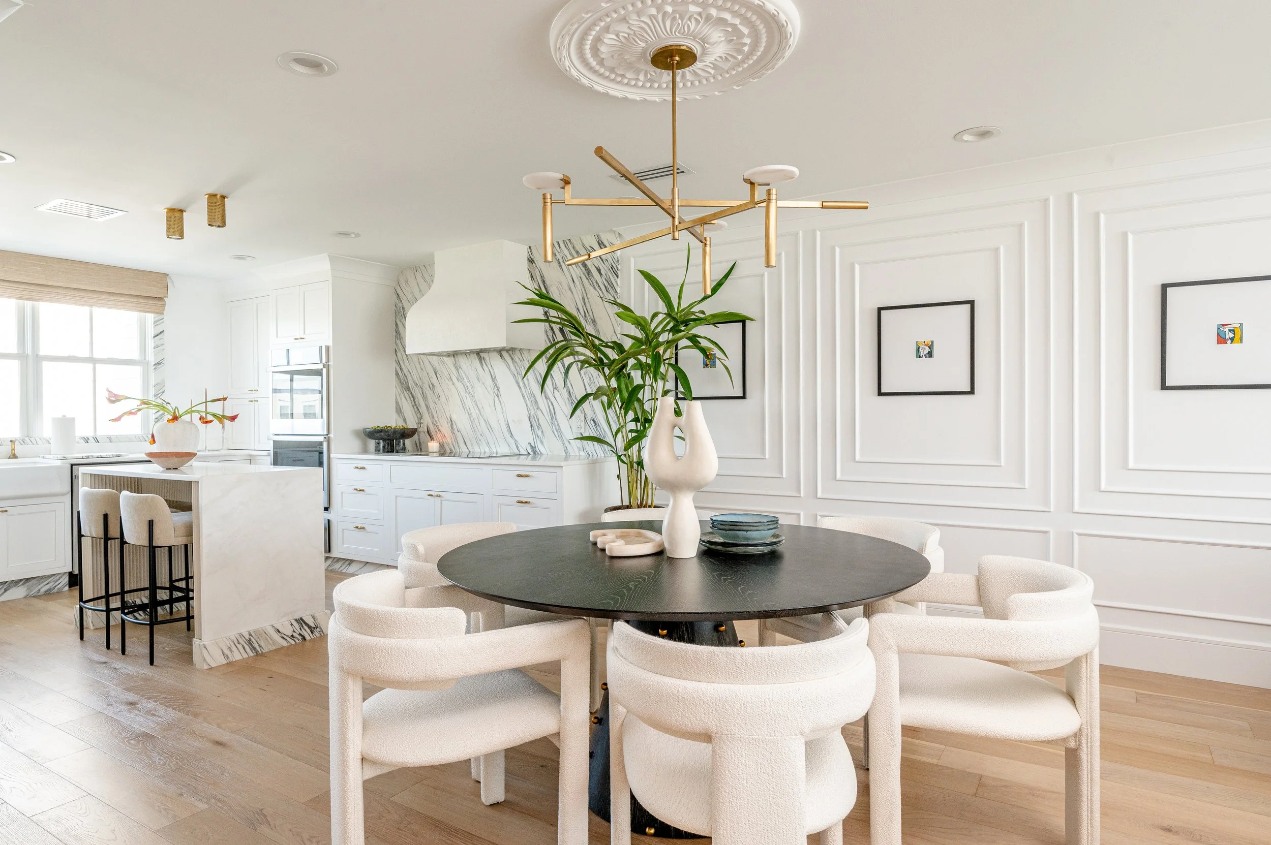 Modern white kitchen and dining area with a black round table, white chairs, a large green plant, and minimalist wall art.