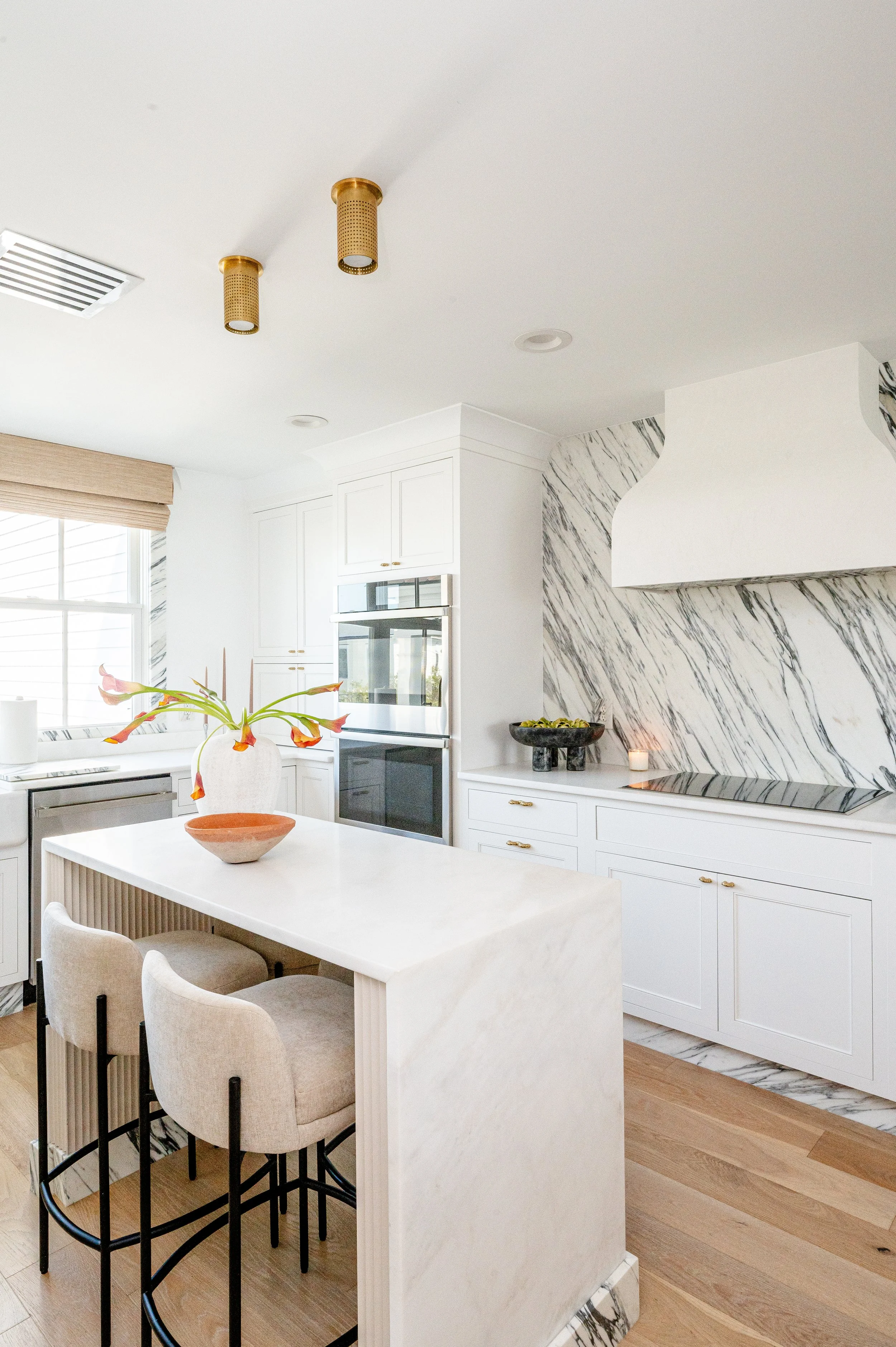 Modern white kitchen with marble backsplash, white cabinets, wooden flooring, and a central island with a vase of orange flowers.