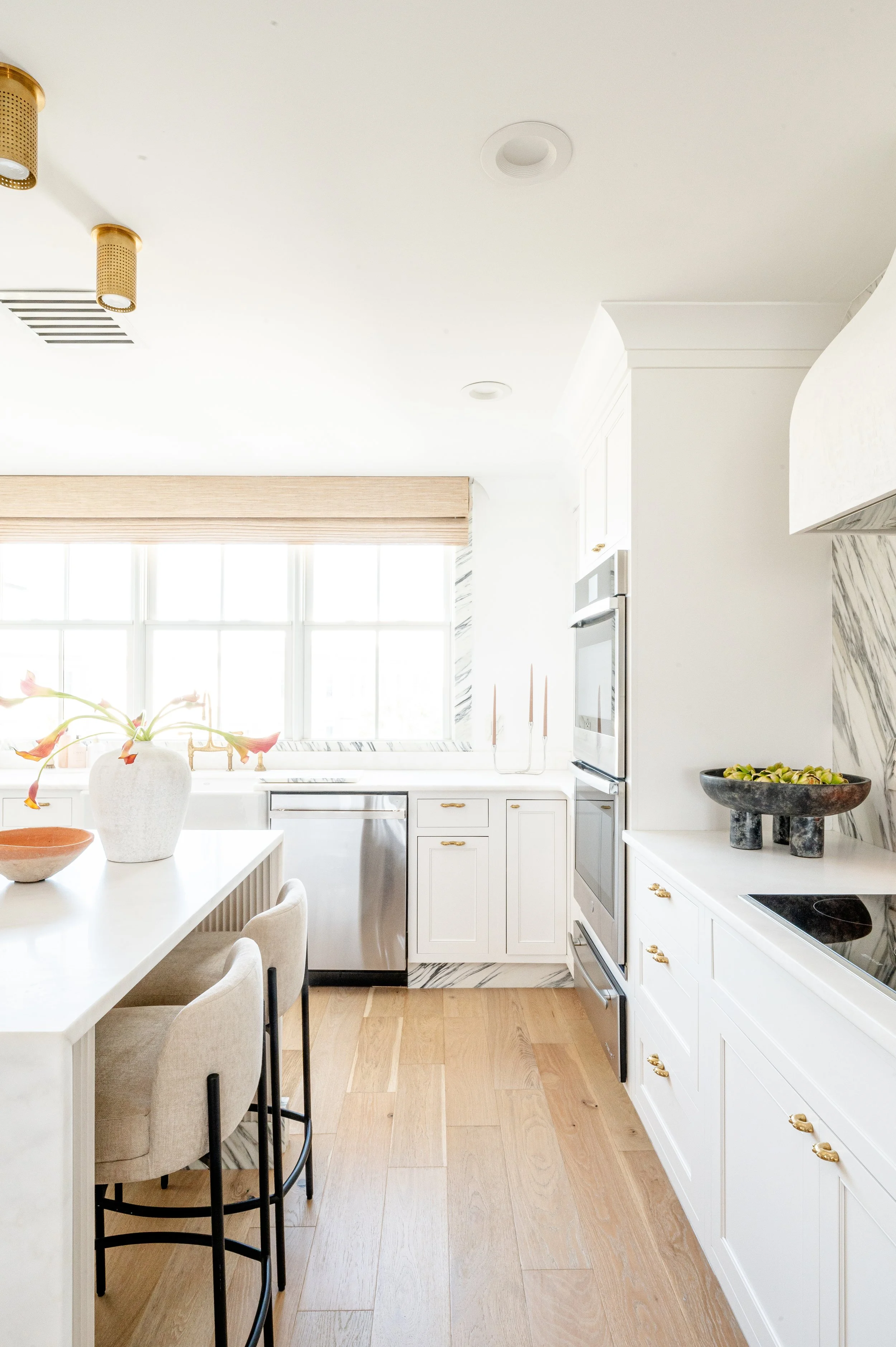 Bright kitchen with white cabinets, a wooden floor, a large window, and modern appliances. Decor includes a white vase with flowers, a bowl, and a black bowl filled with fruit.