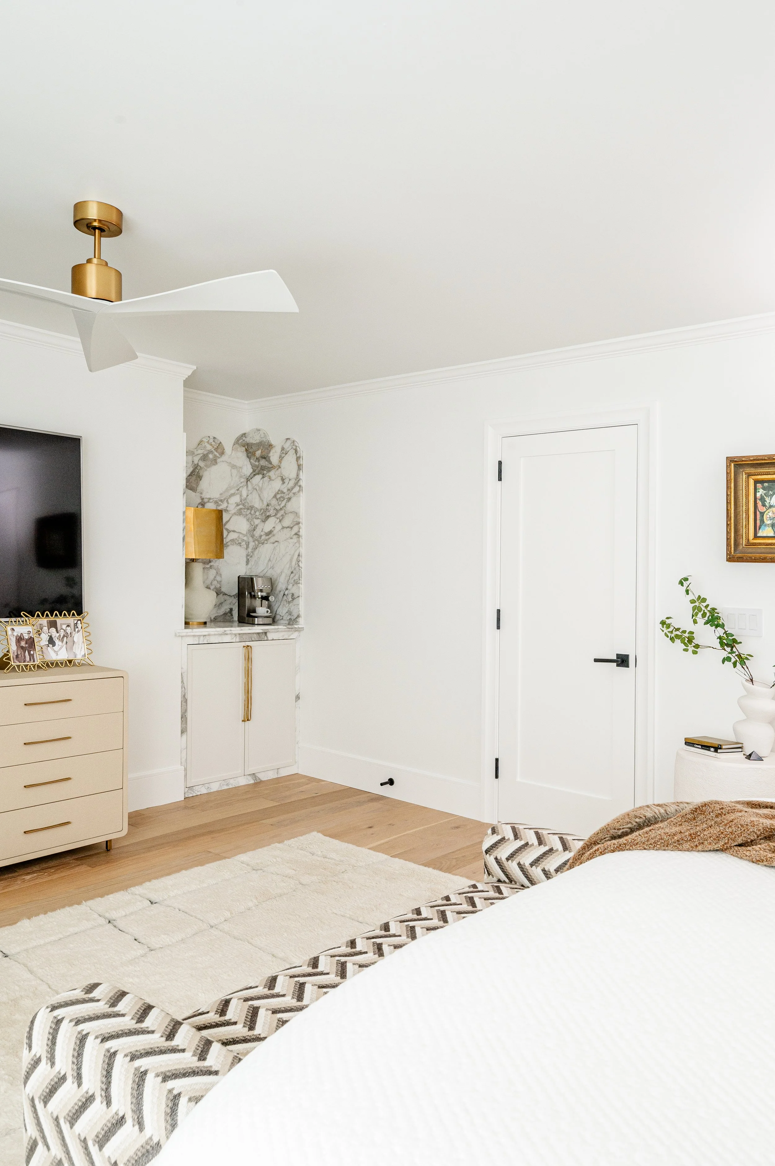 A modern bedroom with white walls and ceiling, a ceiling fan with white blades, a beige dresser with framed photos, a wall-mounted flat-screen TV, a corner marble shelf with a coffee machine, a white door, a side table with a plant, and a bed with a 