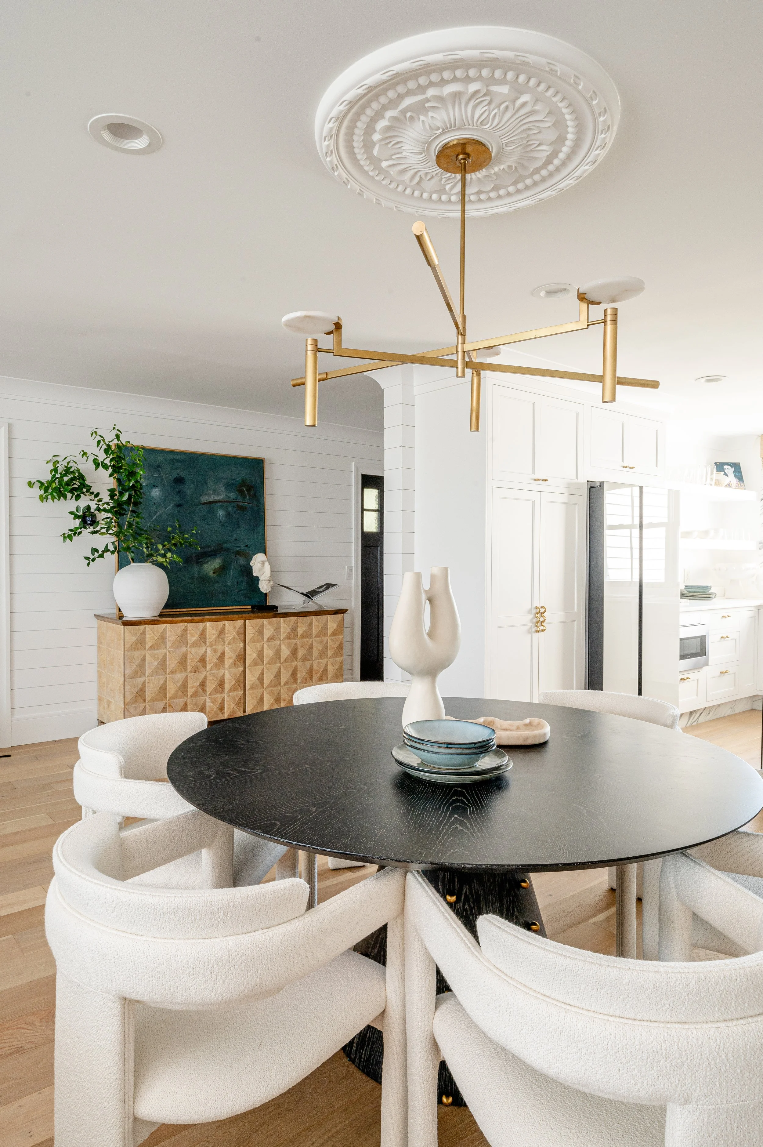 Modern dining room with black oval table, six white upholstered chairs, decorative vases, and a gold-toned chandelier