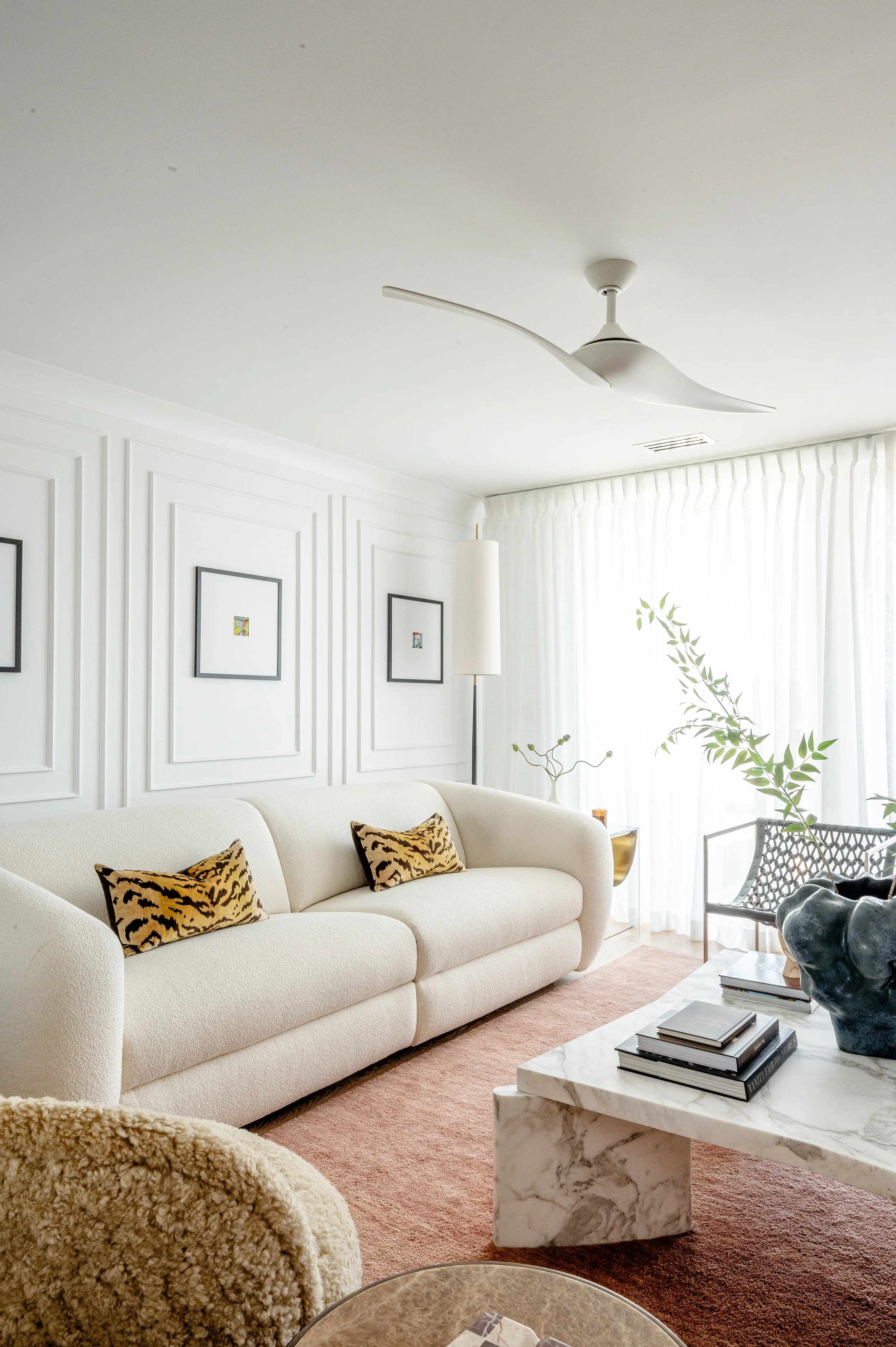 A modern living room with a white sofa decorated with animal print pillows, a marble coffee table, green plants, a white wall with framed artwork, a white ceiling fan, and sheer white curtains letting in natural light.