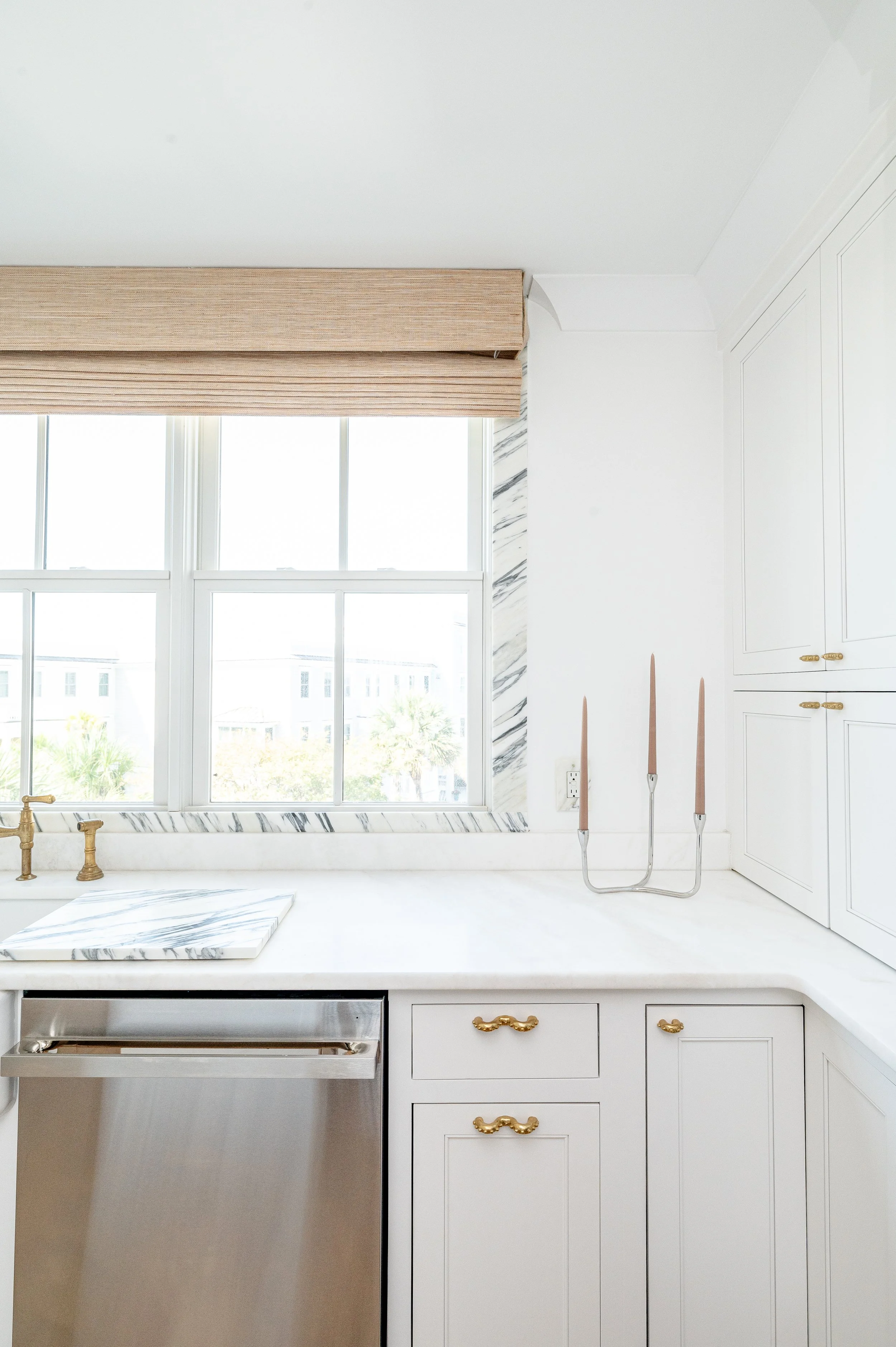 Bright white kitchen with large window, marble backsplash, and gold hardware on cabinets.
