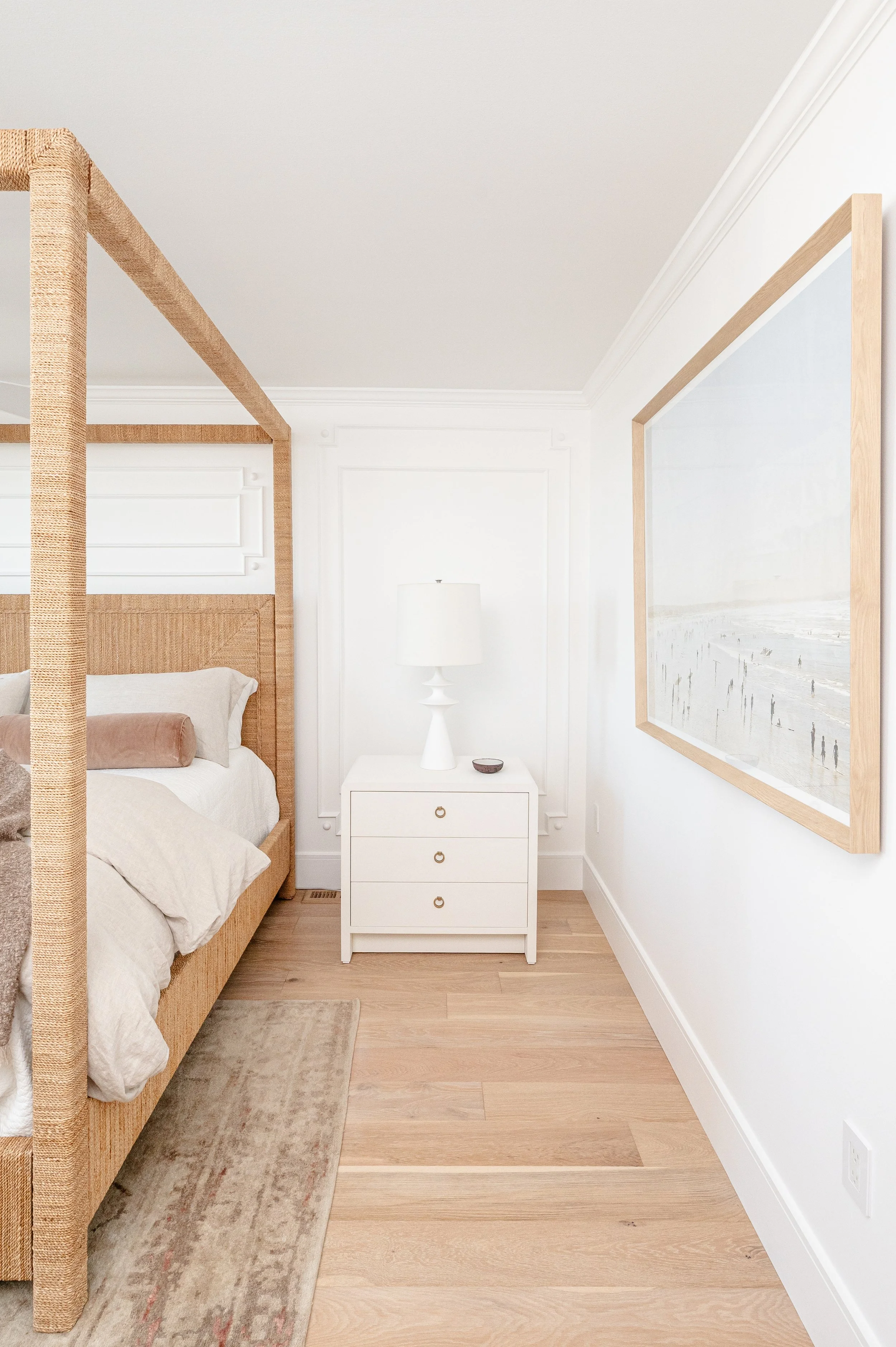A bedroom corner with a white nightstand, a white table lamp, a beige bed with pillows, a framed artwork on the white wall, and wooden flooring.