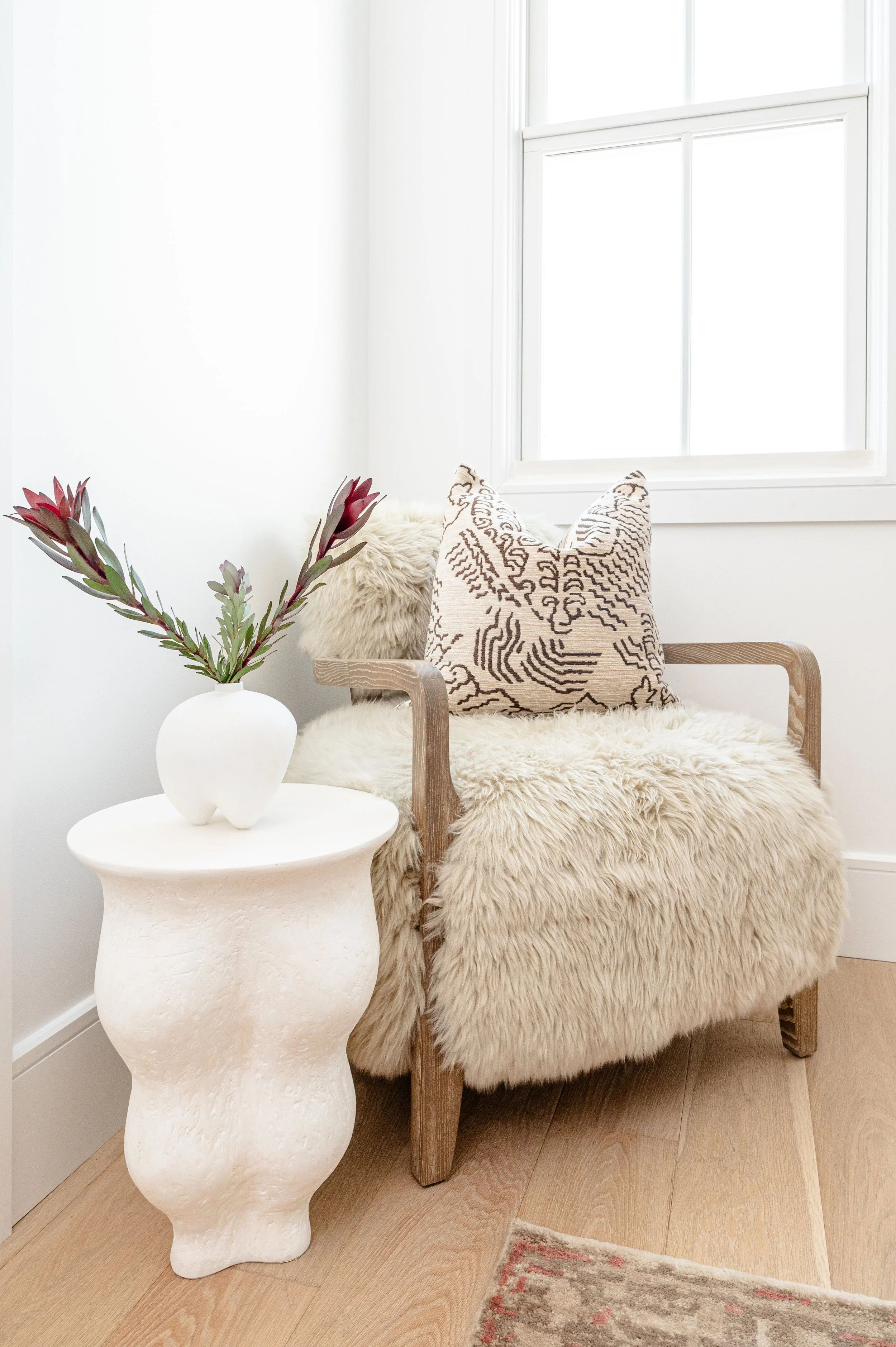 A cozy corner with a beige fur-covered armchair, a decorative cushion, a white side table with a white vase holding red-tipped green leaves, by a window with white trim.